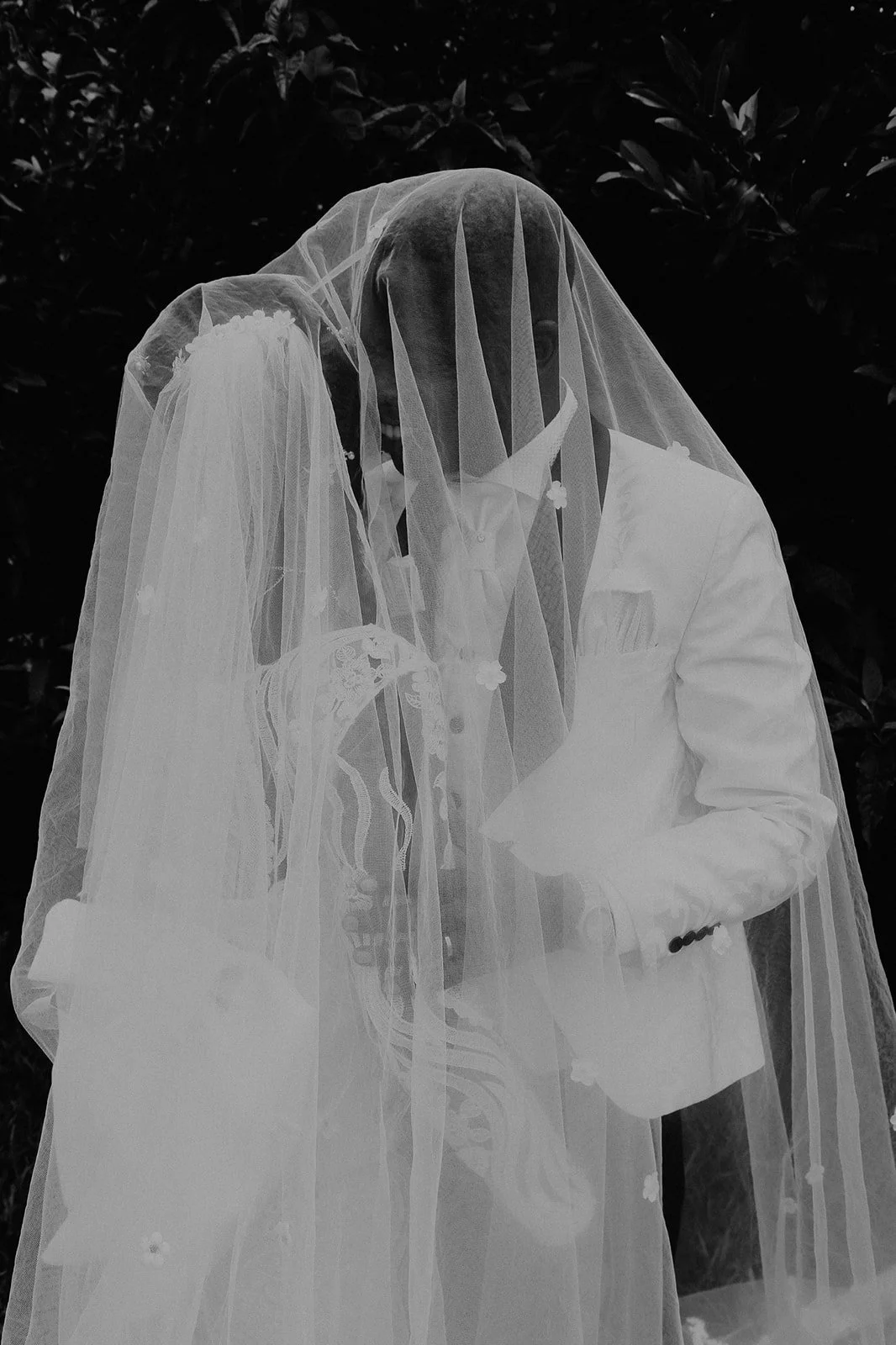 A black-and-white photograph of a bride and groom standing close together. The bride is wearing a veil and dress with intricate embroidery, while the groom is in a white suit. The bride is holding a small bouquet, and the groom is reading from a piec