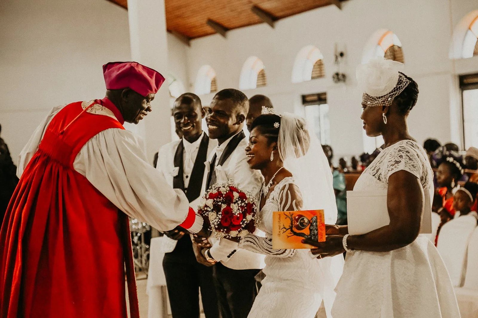 A wedding ceremony with a bride and groom exchanging vows in a church, surrounded by witnesses and family members, with the officiant in a red robe standing in front of them.