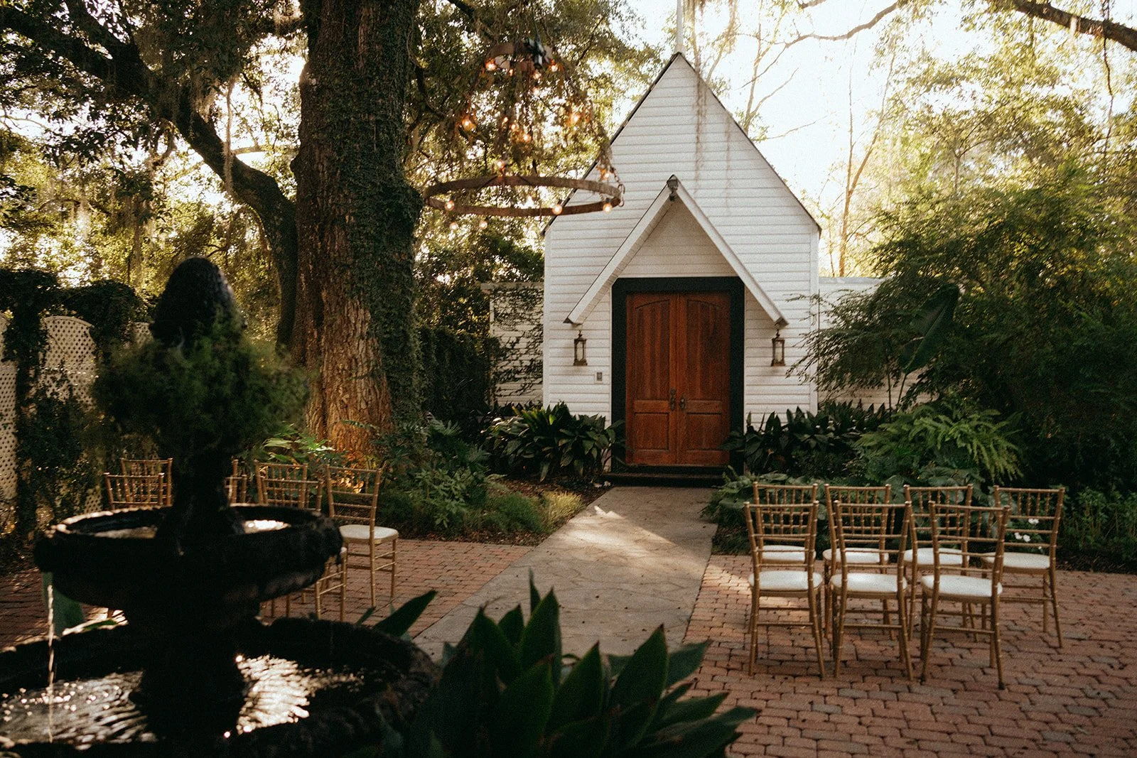 Outdoor wedding ceremony setup with rows of chairs, a small white chapel with brown doors, greenery, trees, and a fountain in the foreground.
