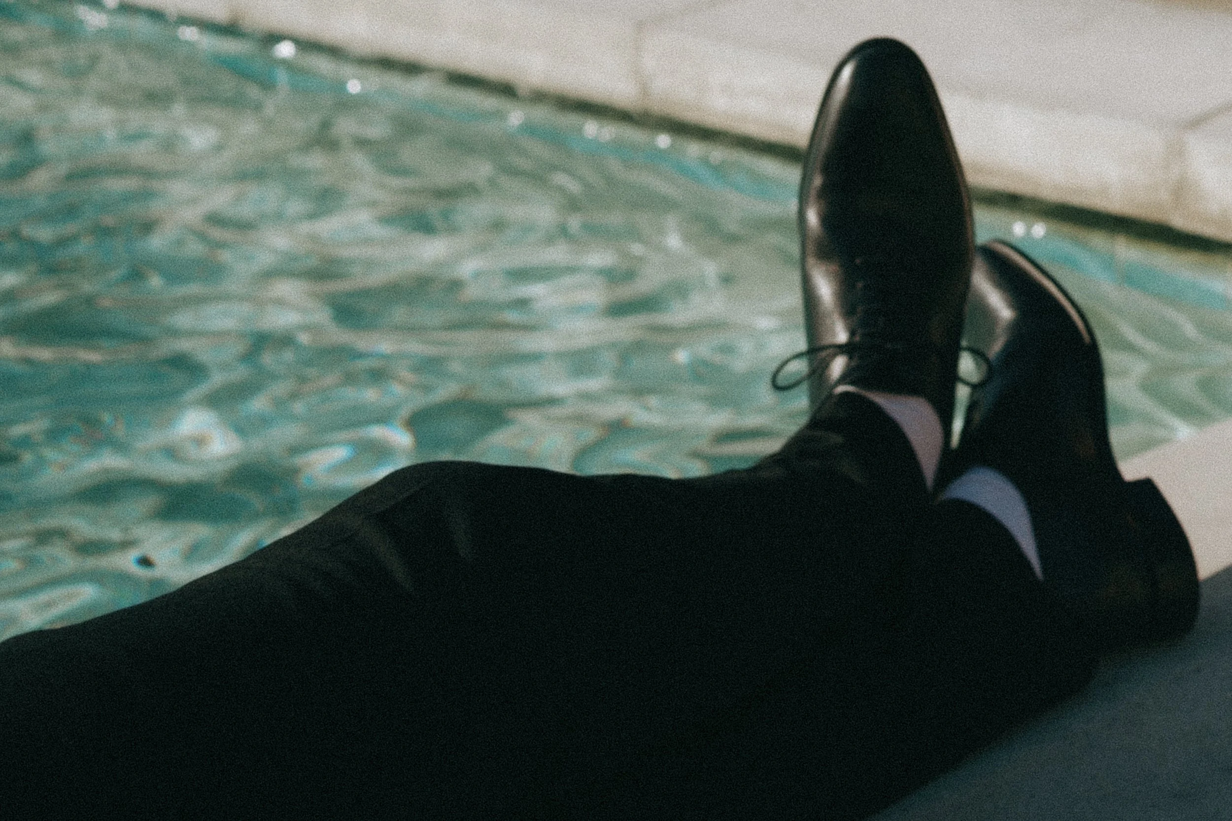 Person in black dress shoes and black pants sitting near a swimming pool with their legs stretched out.