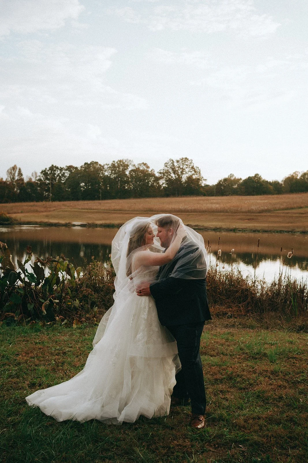 A bride and groom embrace by a pond in a rural outdoor setting, with trees and open fields in the background.