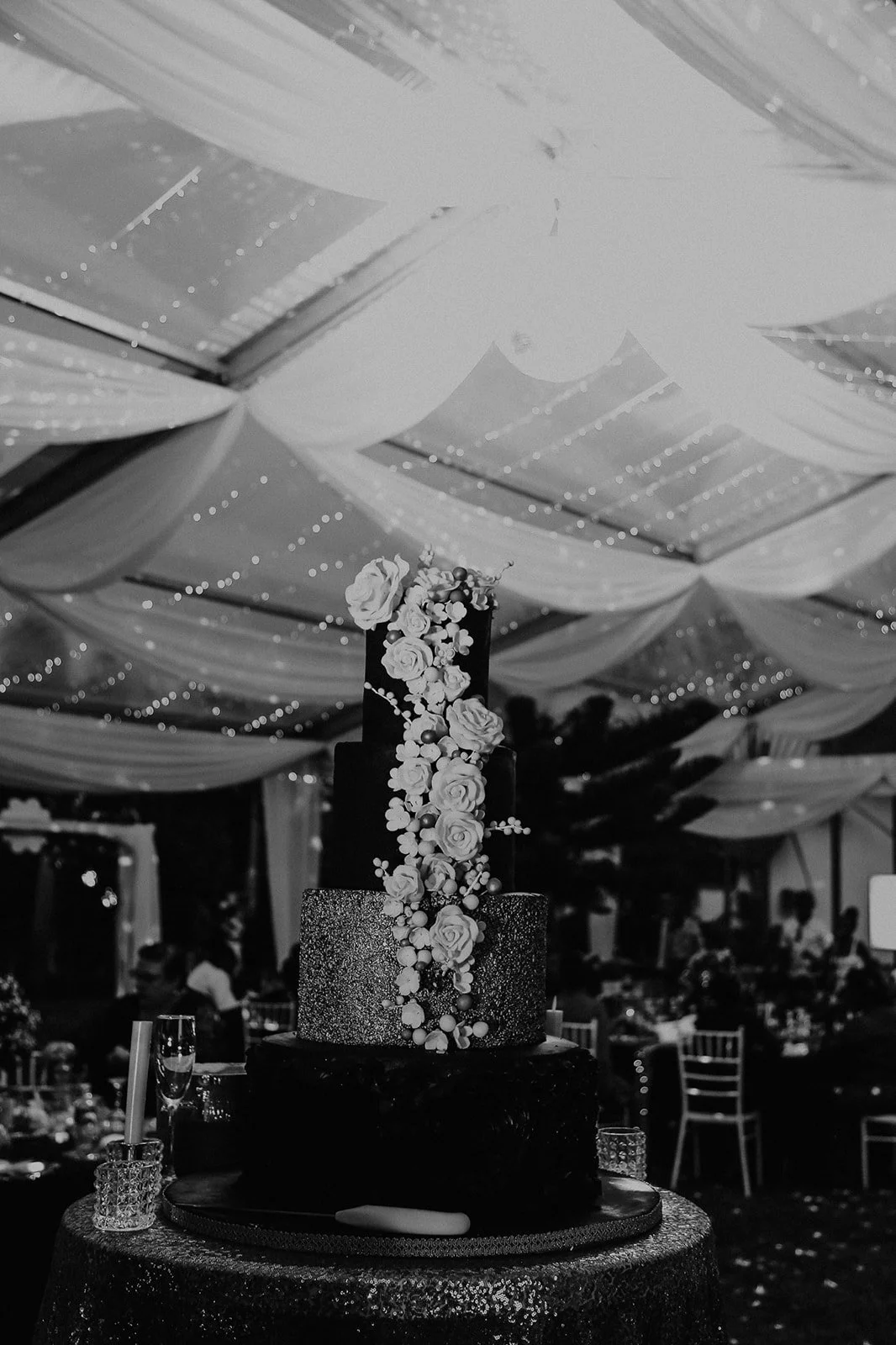 A three-tiered wedding cake decorated with roses and berries, placed on a table with champagne glasses in a decorated reception tent with draped fabric and string lights.