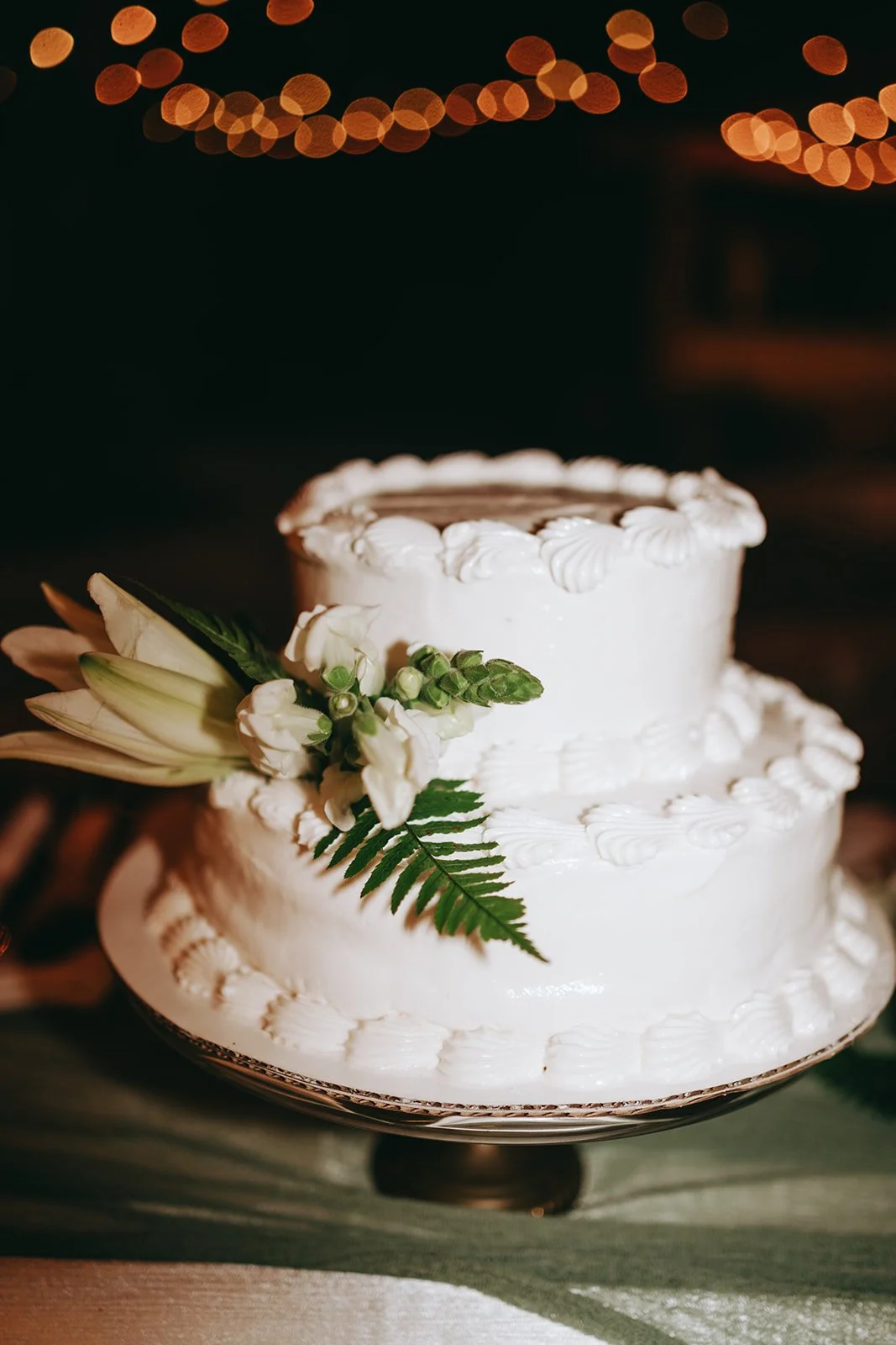 Two-tiered white wedding cake decorated with flower and greenery, with a gold cake stand, set against a dark background with blurred orange string lights.