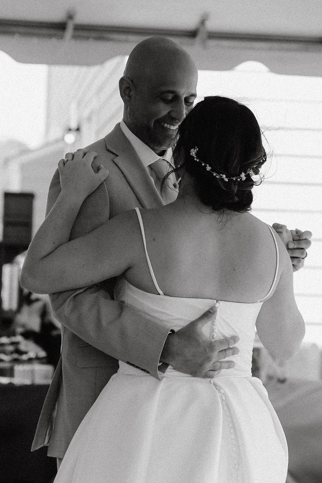 A black-and-white photo of a couple dancing closely, smiling, with the man in a light-colored suit and the woman in a wedding dress with a floral hairpiece, in a room with a window.