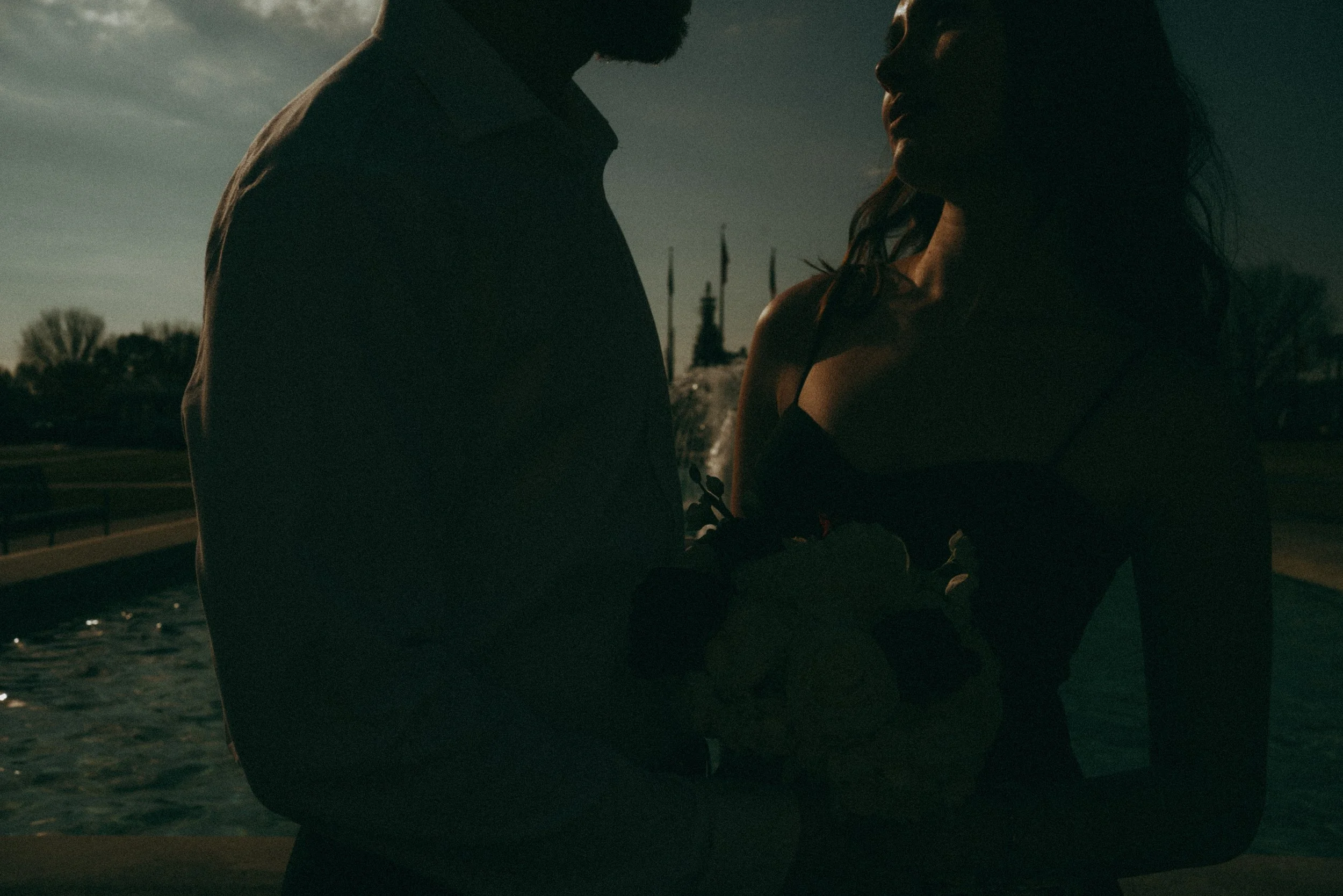 Silhouettes of a man and woman holding a baby near a fountain at dusk.