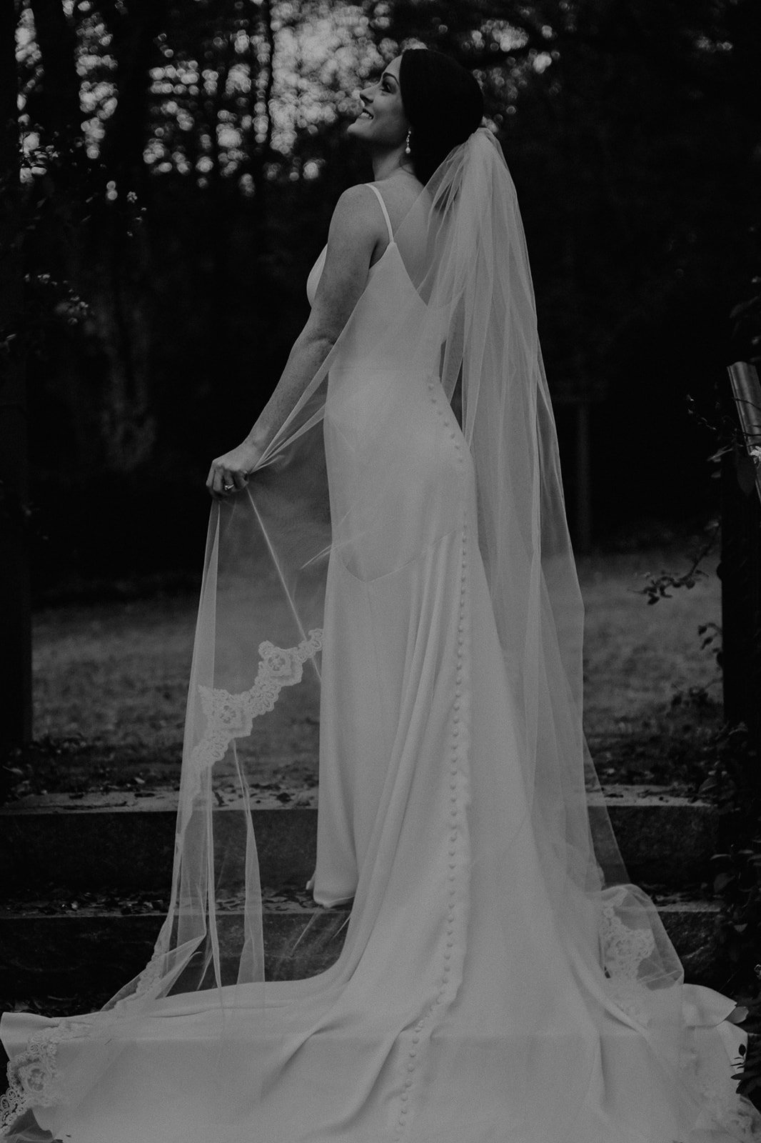 A woman in a wedding dress and veil standing outdoors, smiling, in a black and white photo.