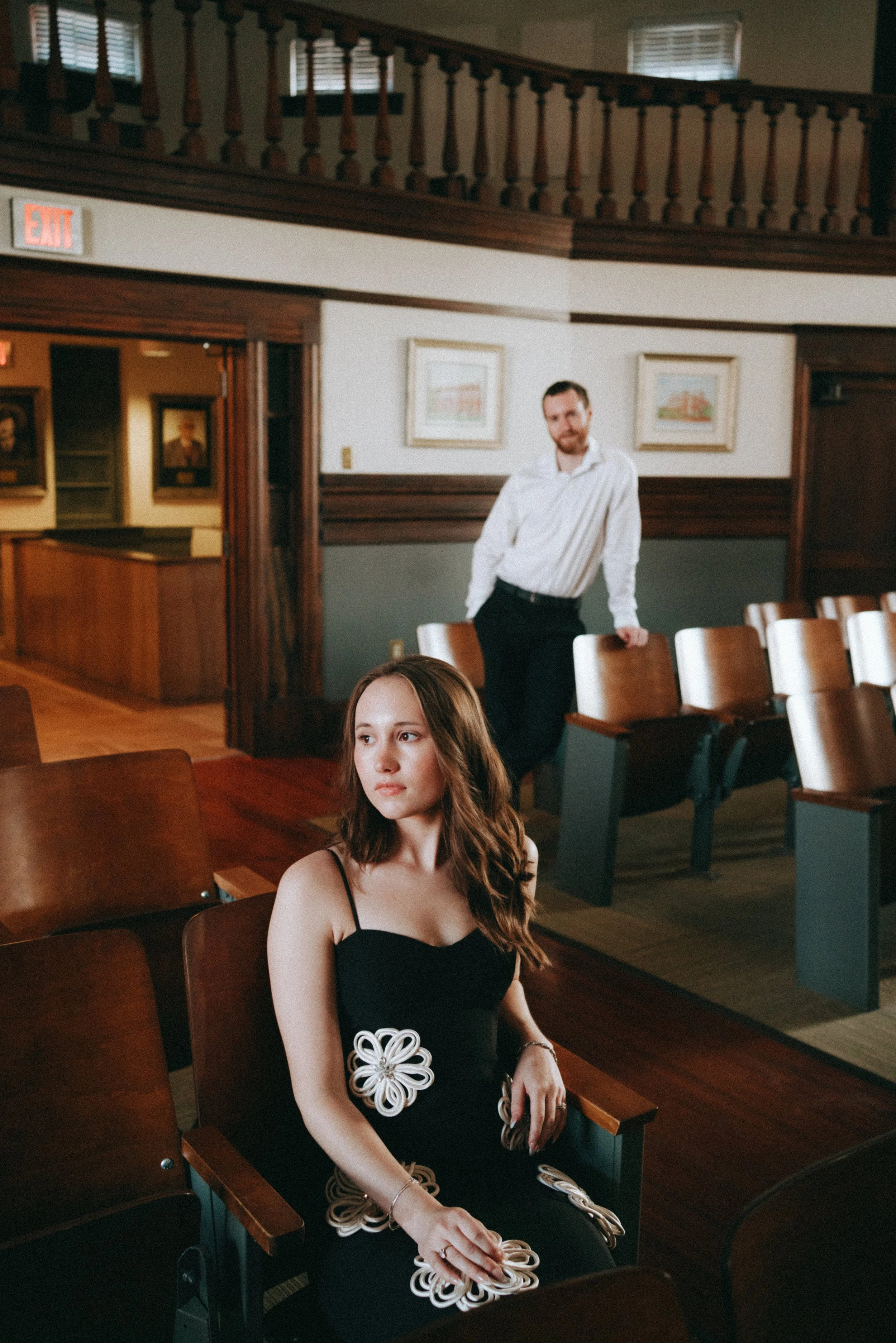 A young woman in a black dress with flower-shaped embellishments sitting in a wooden auditorium seat, with a man in a white shirt standing behind her in a room with framed pictures on the wall.
