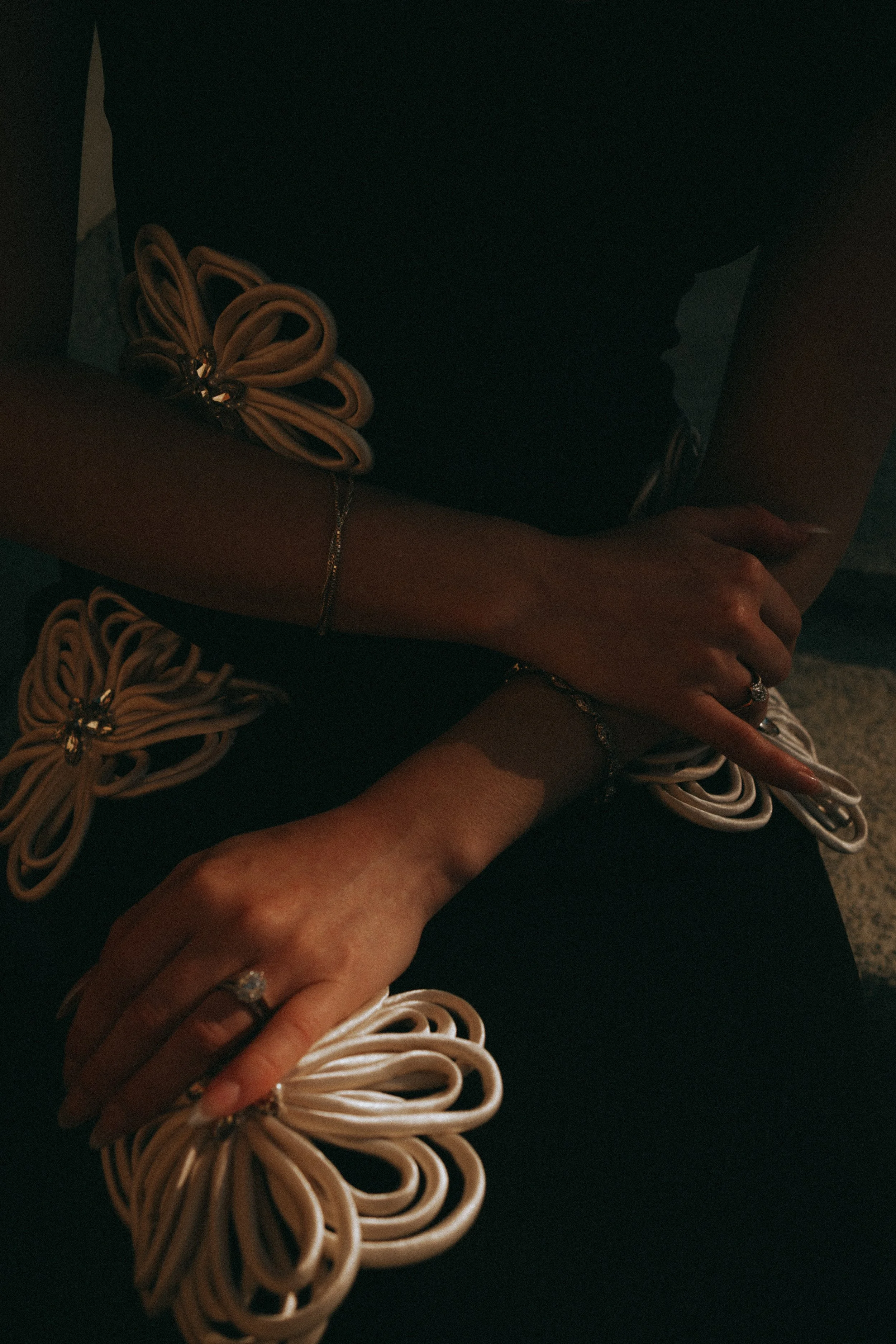 Close-up of a person's hands resting on their lap, wearing rings and bracelets, with decorative flower-shaped embellishments on a dress or outfit.