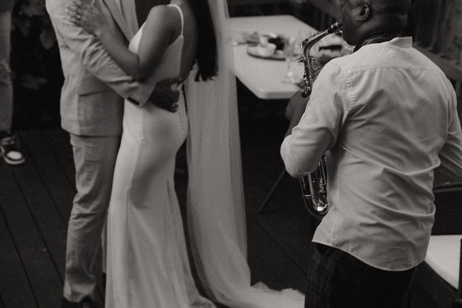 A man playing a saxophone at a social event, with a woman holding a drink in the background.