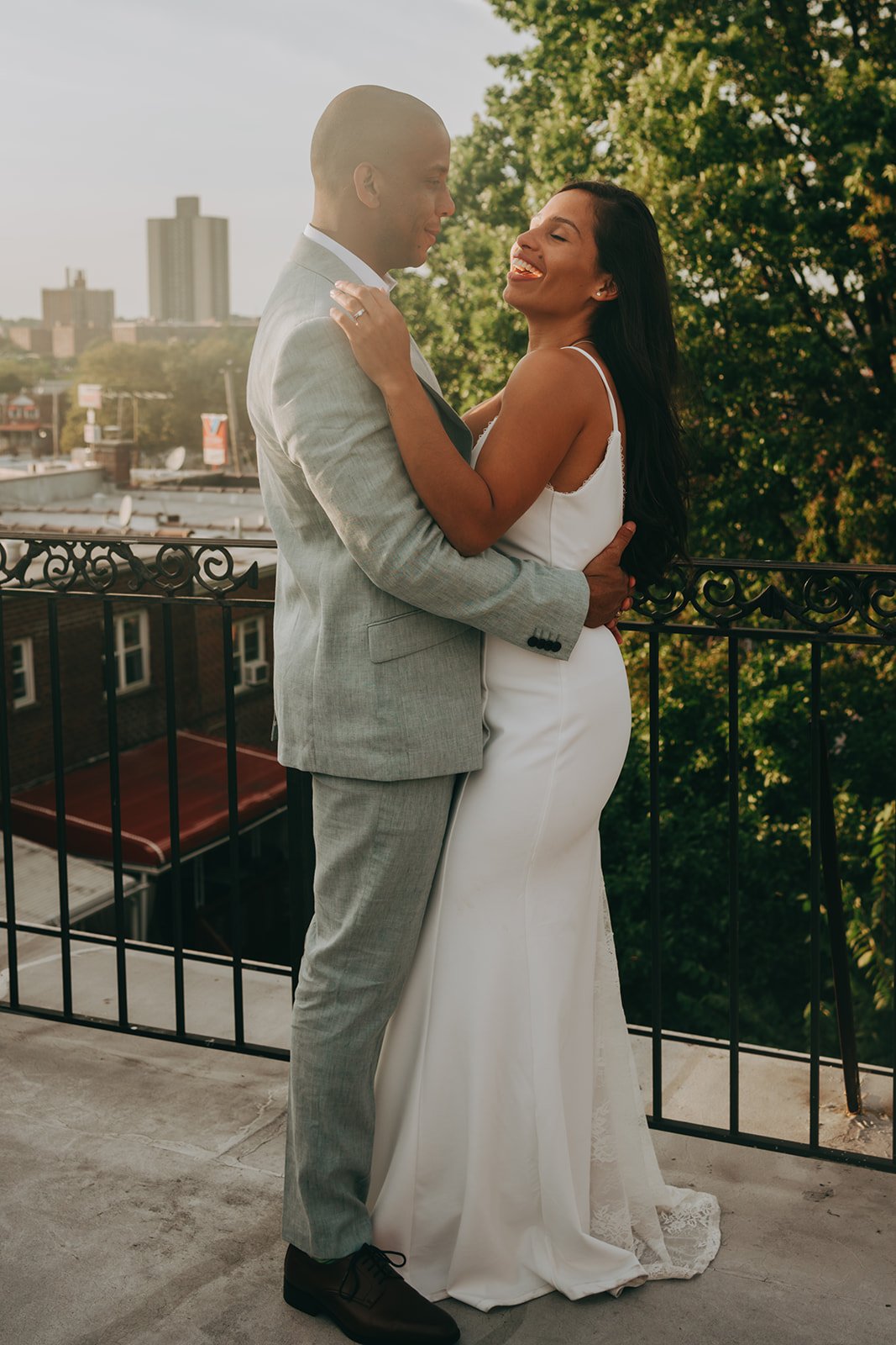 A couple in wedding attire embracing on a balcony, with trees and city buildings in the background.