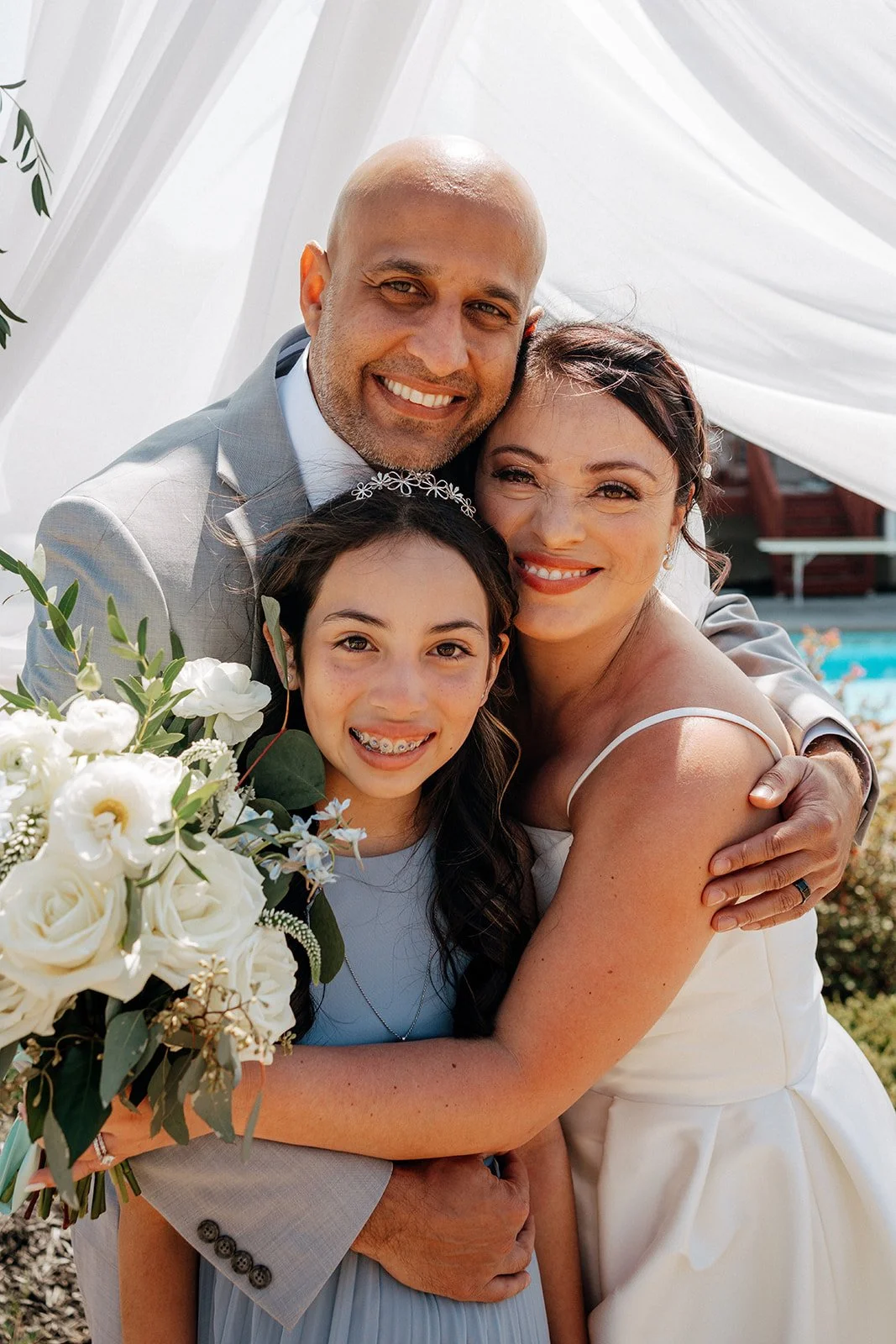 A wedding photo of a joyful couple and a young girl, all smiling and hugging, outdoors with a white canopy and a pool in the background. The bride is wearing a white wedding dress, the groom in a light grey suit, and the girl in a light blue dress wi