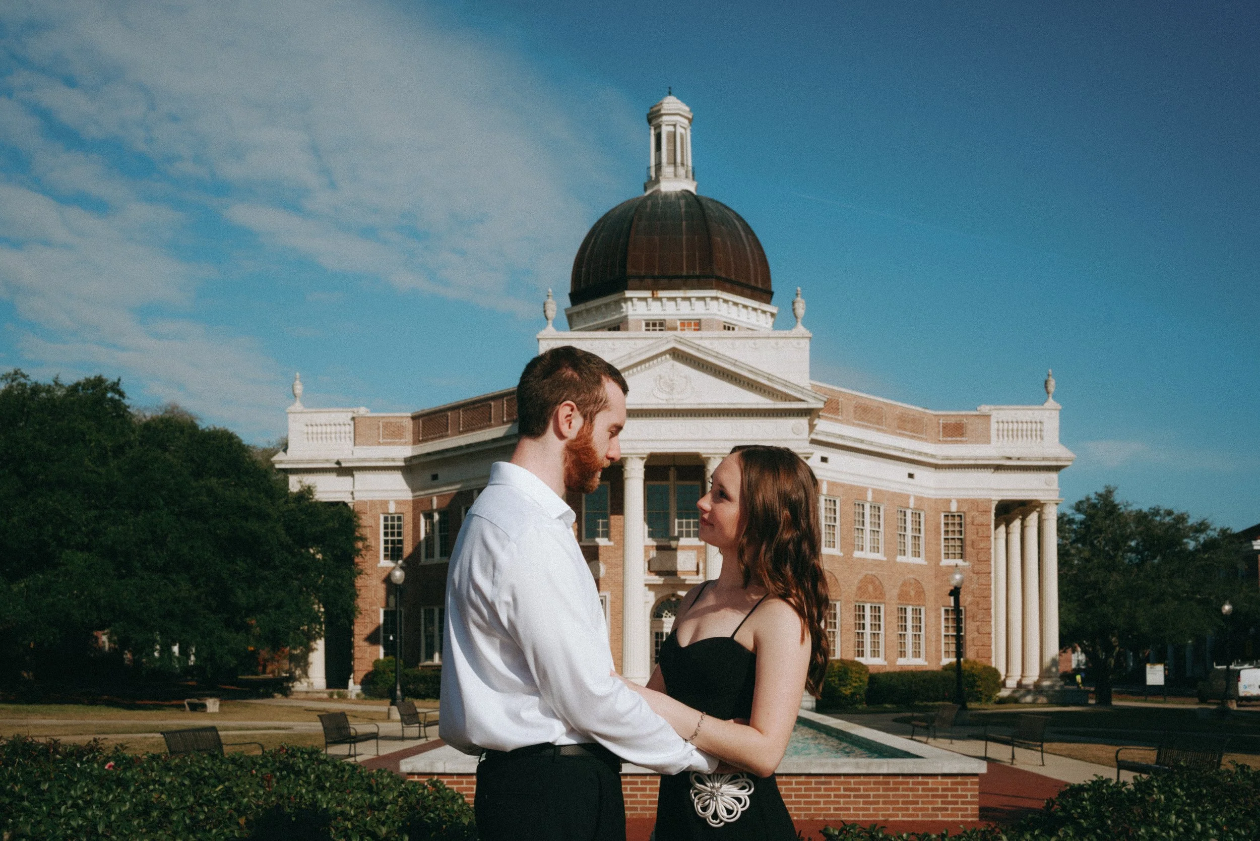 A couple is standing in front of a historic building with a large dome, holding each other's hands and looking into each other's eyes on a sunny day.