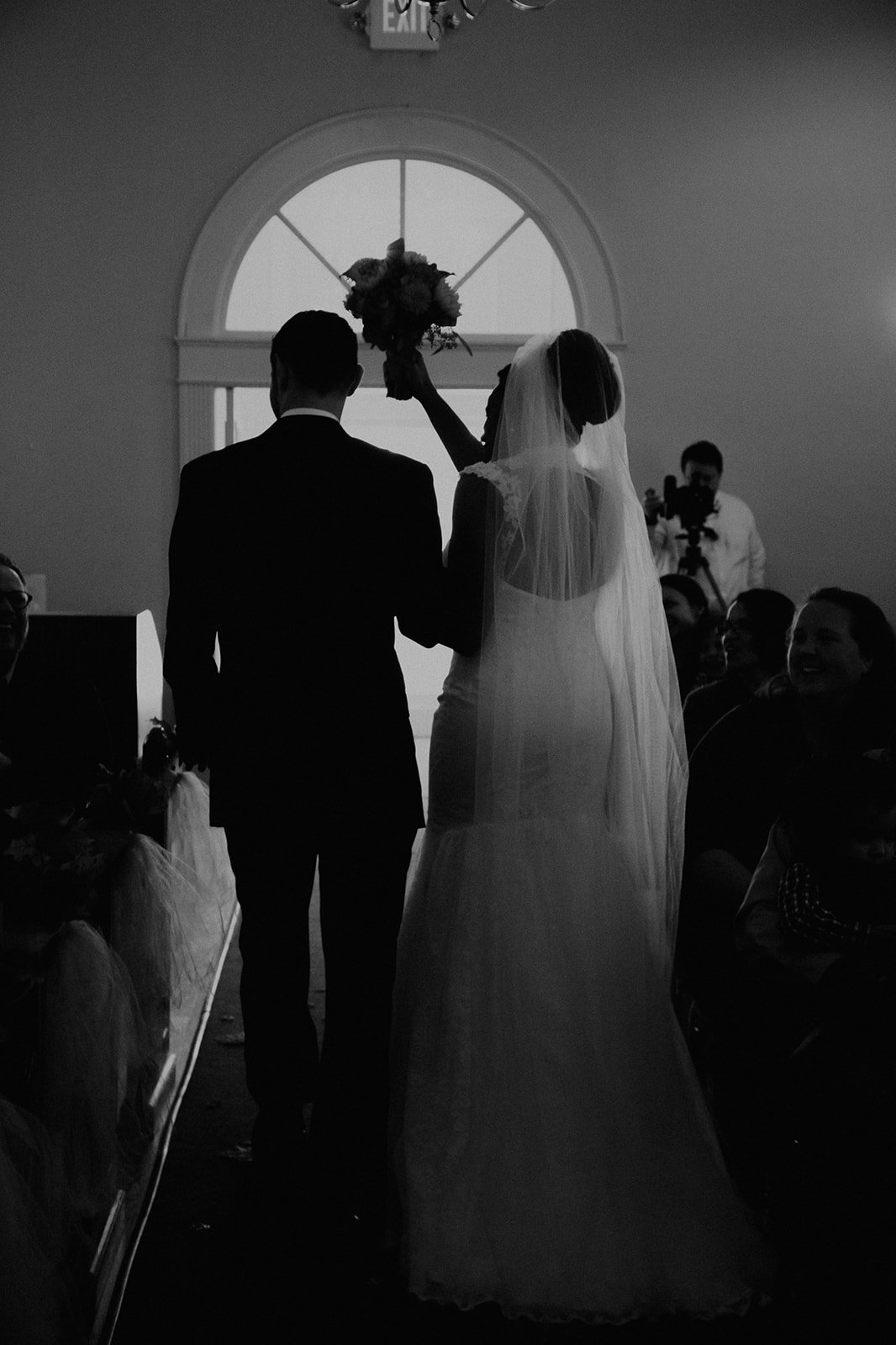 Silhouette of a bride and groom during their wedding ceremony with a person holding a bouquet above them, illuminated by backlighting at a church.