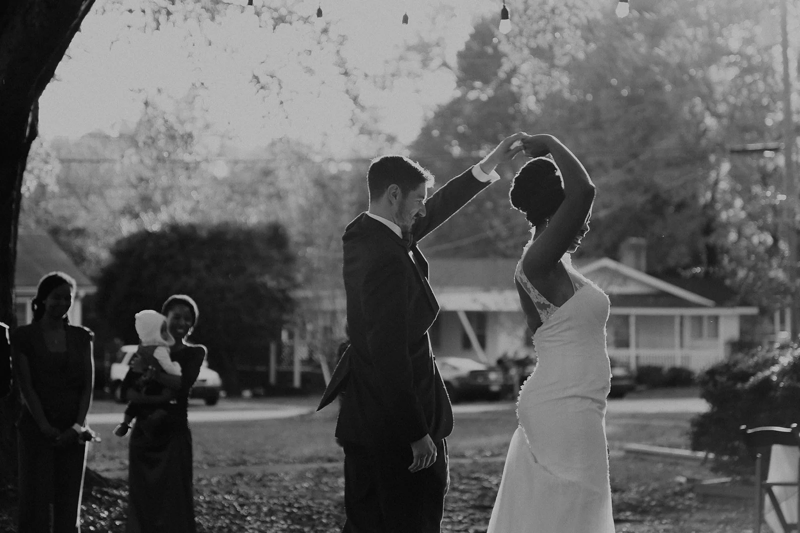 A black and white photo of a couple dancing outdoors during a wedding, with guests in the background, and trees and houses visible in the distance.
