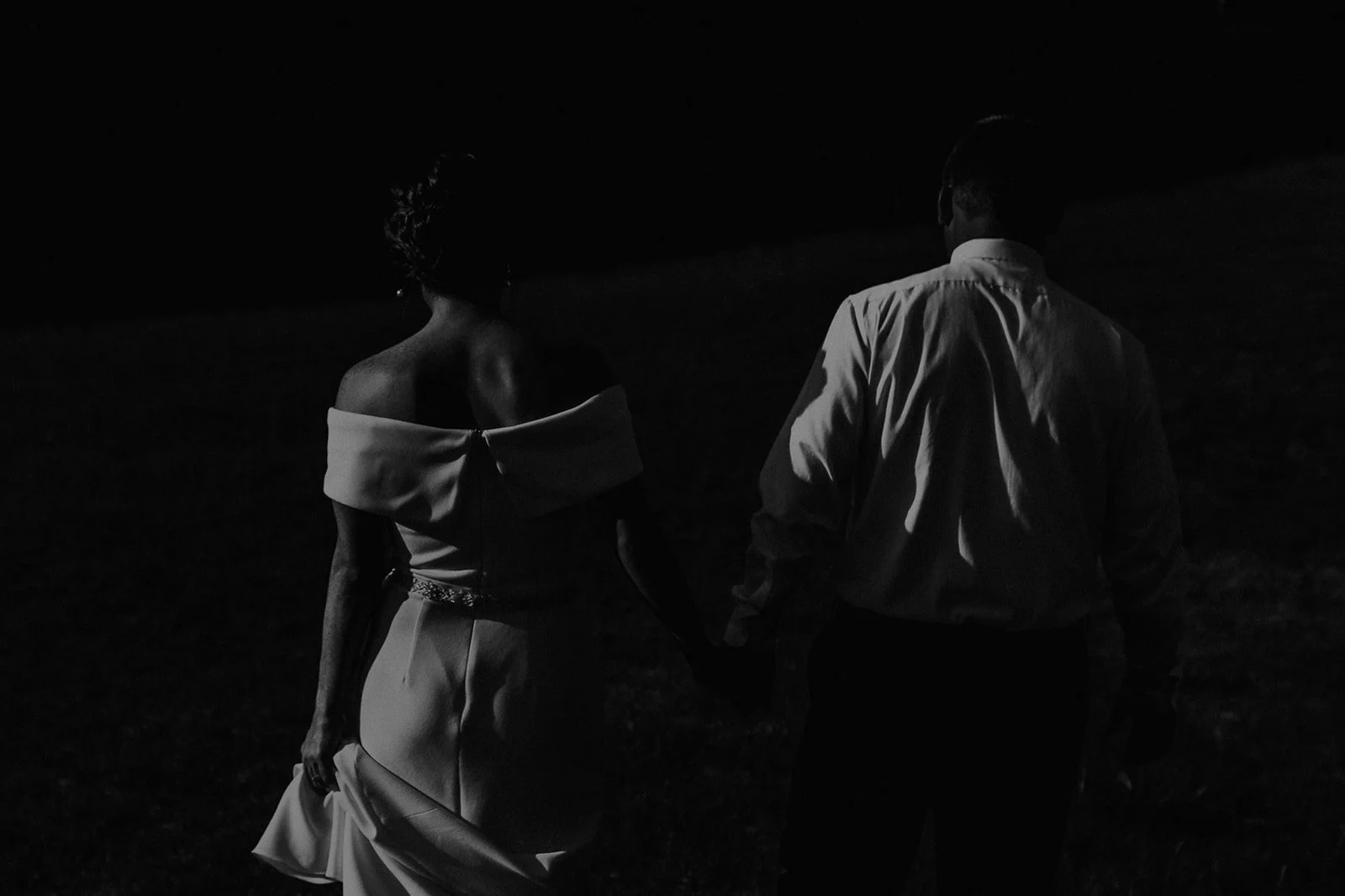 A couple holding hands at night, with the woman wearing an off-shoulder dress and the man dressed in a shirt and pants.