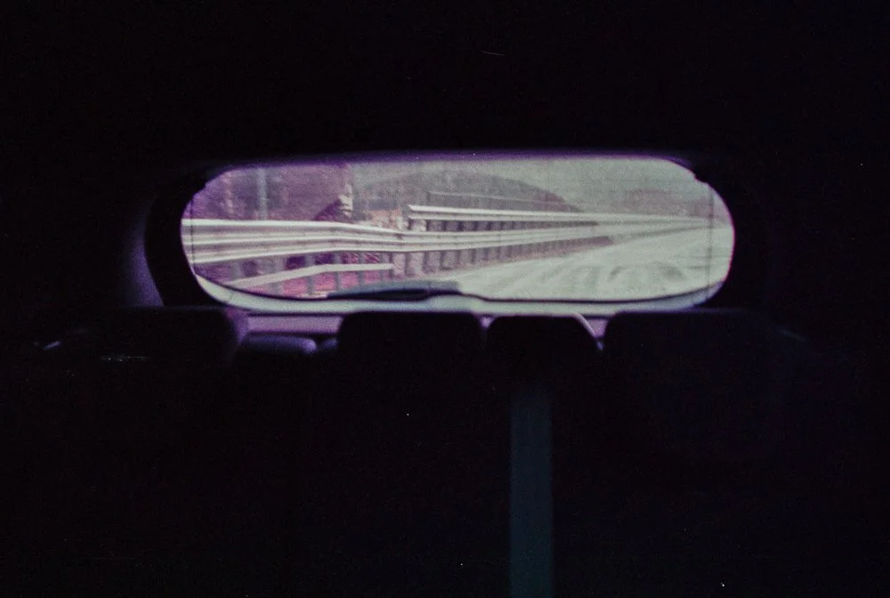 Vista della strada attraverso il lunotto di un'auto, con parapetti laterali visibili.