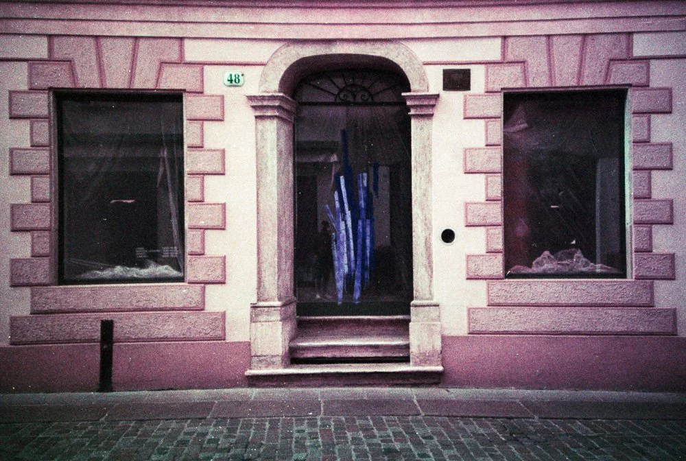 Facciata di edificio con ingresso centrale e vetrine laterali su strada acciottolata, tonalità rosa e viola.