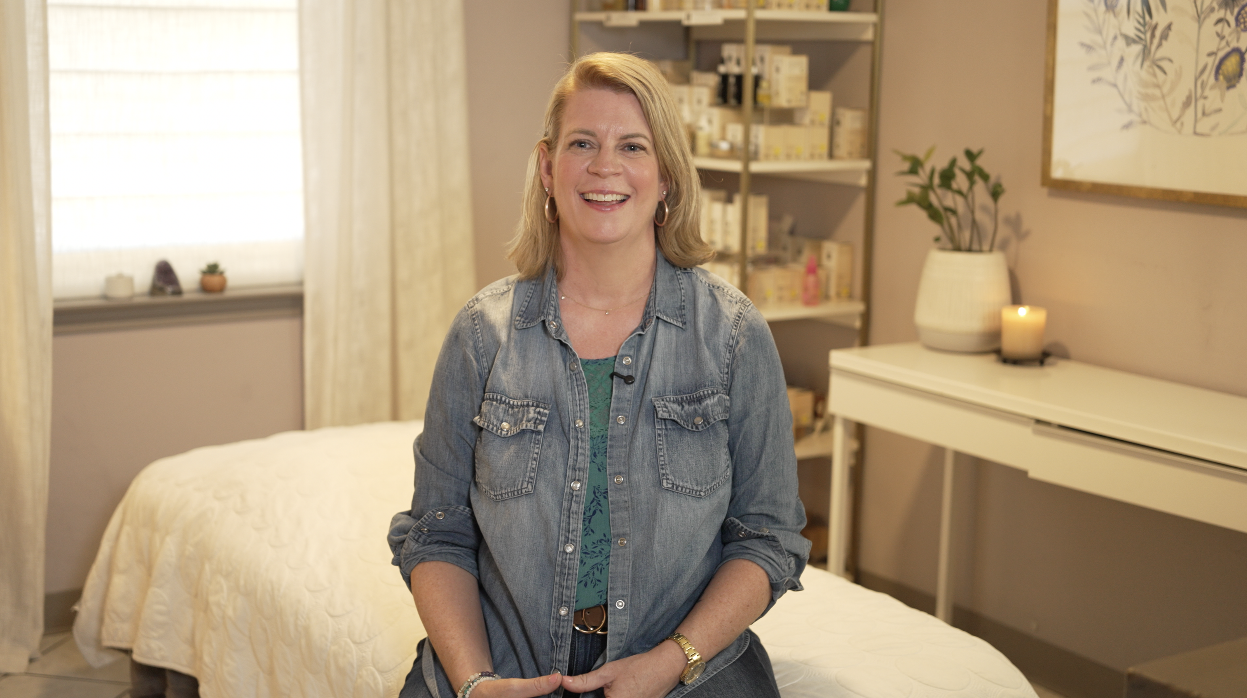 A woman with blonde hair, wearing a denim jacket and jewelry, sitting on a bed in a cozy bedroom with light-colored walls, a white desk, a potted plant, a lit candle, and a bookshelf in the background.