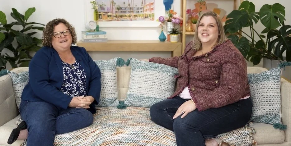 Two women sitting on a colorful couch in a brightly decorated room, smiling at the camera.
