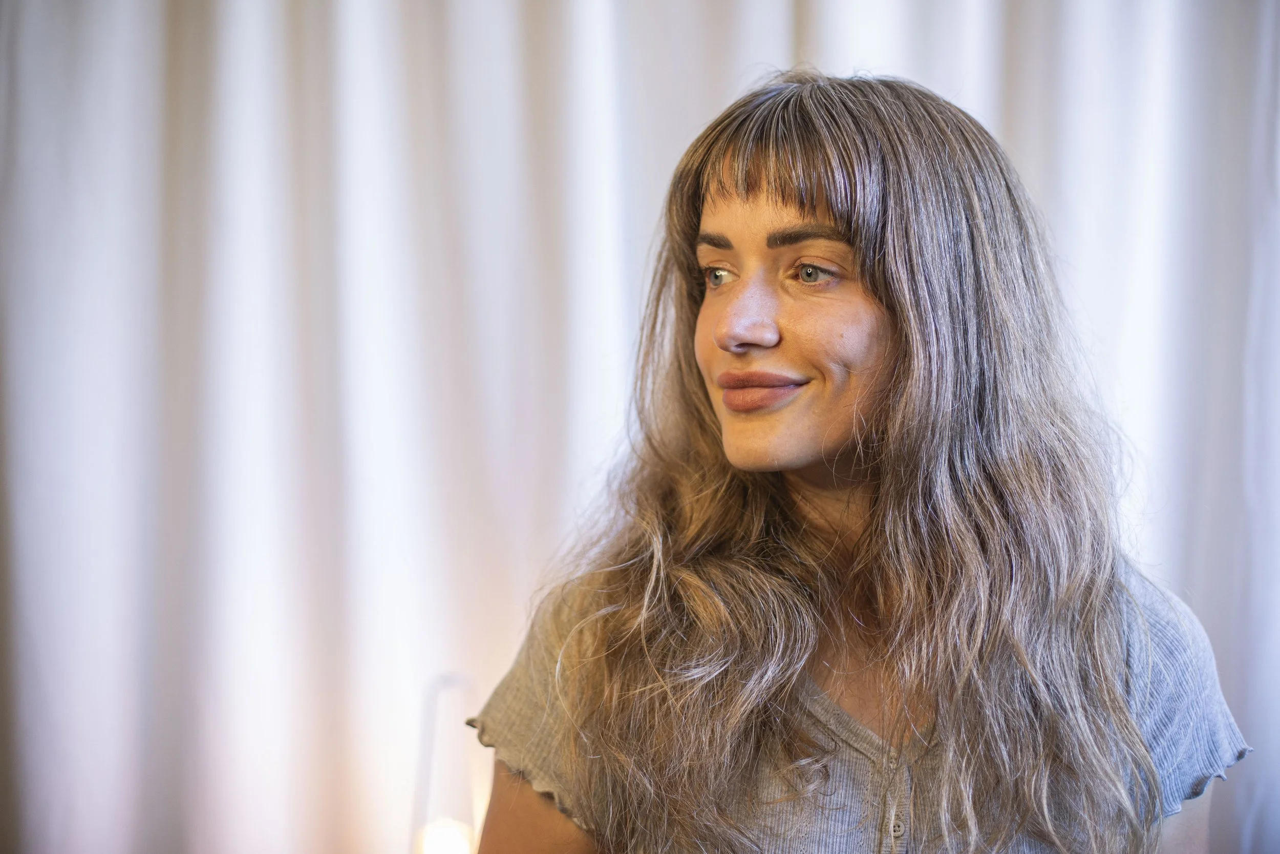 Portrait of a woman with long wavy hair and bangs, smiling softly, in front of a background of cream-colored curtains.