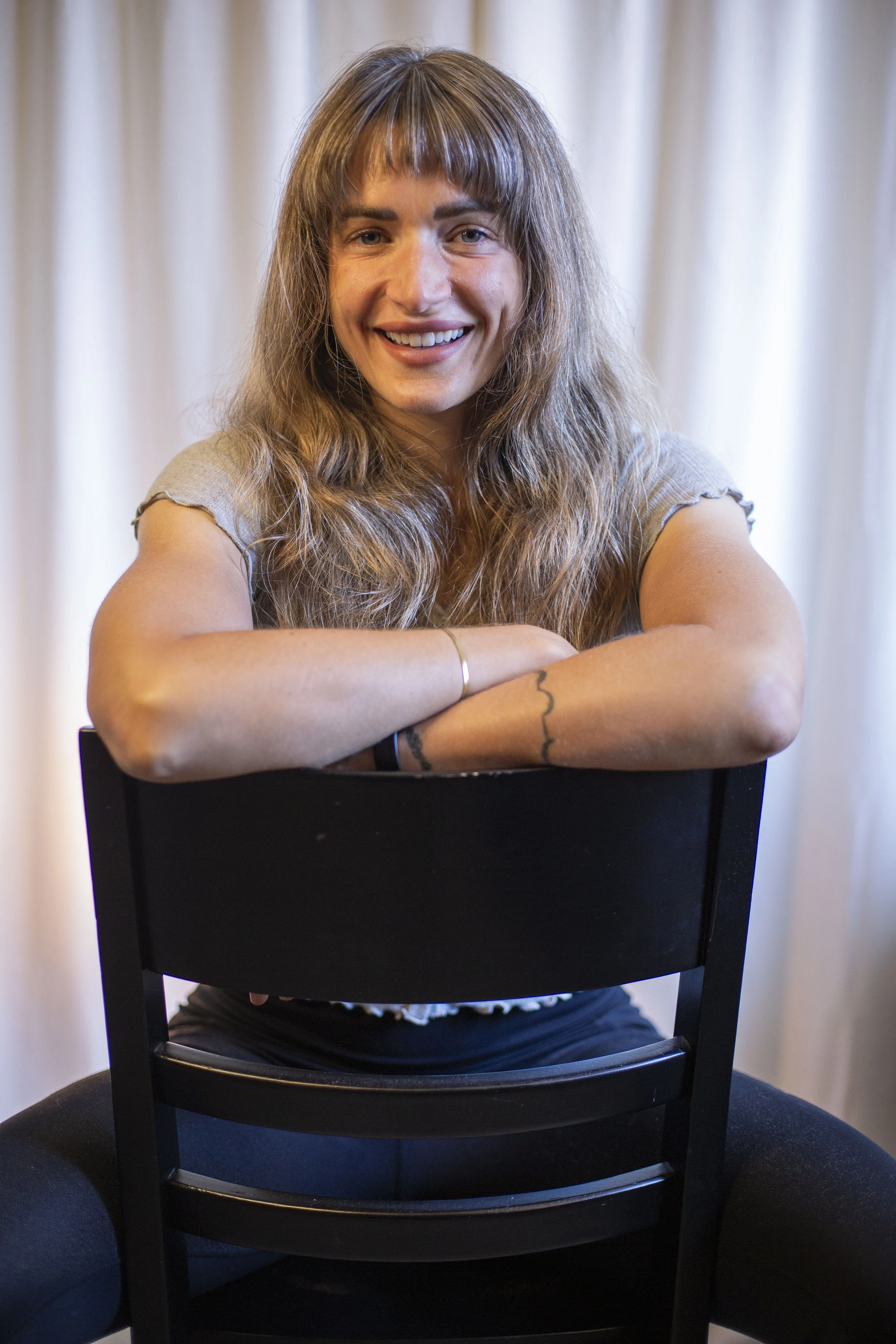 A young woman with long, wavy brown hair and a wide smile, sitting behind a black chair with her arms resting on the top of the chair back. She is wearing a gray short-sleeved top and black pants, with a tattoo on her left forearm and a bracelet on her right wrist. The background features beige curtains.