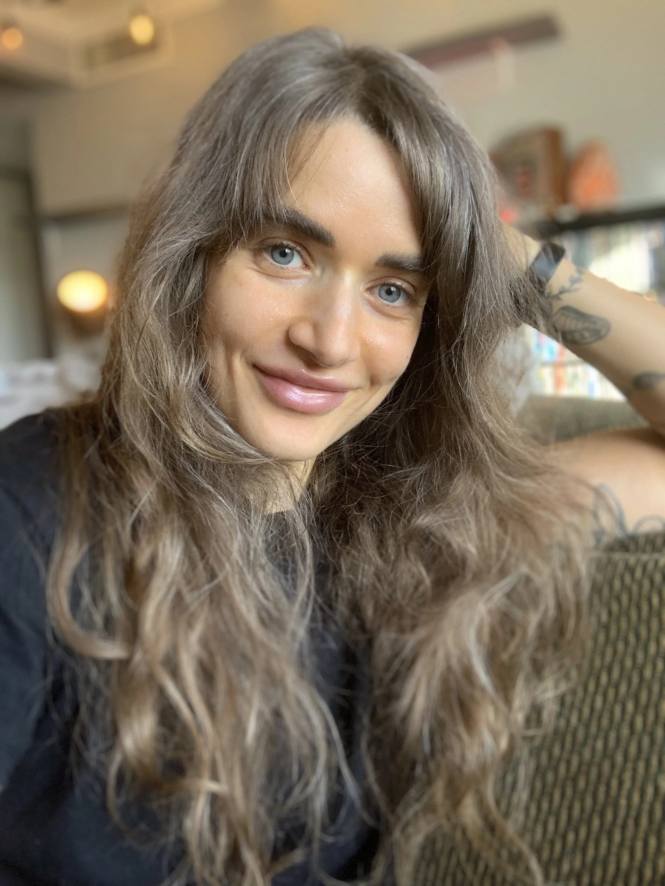 Close-up of a woman with long, wavy brown hair, blue eyes, and a tattoo on her left arm, smiling in a cozy indoor setting.
