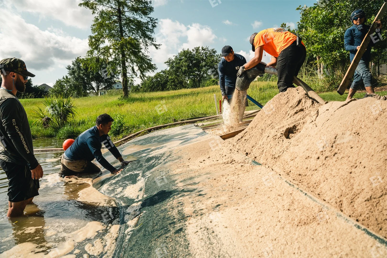Group of people working together to place a large fabric or tarp into a body of water, filling it with sand or soil outside in a grassy area with trees under a partly cloudy sky.