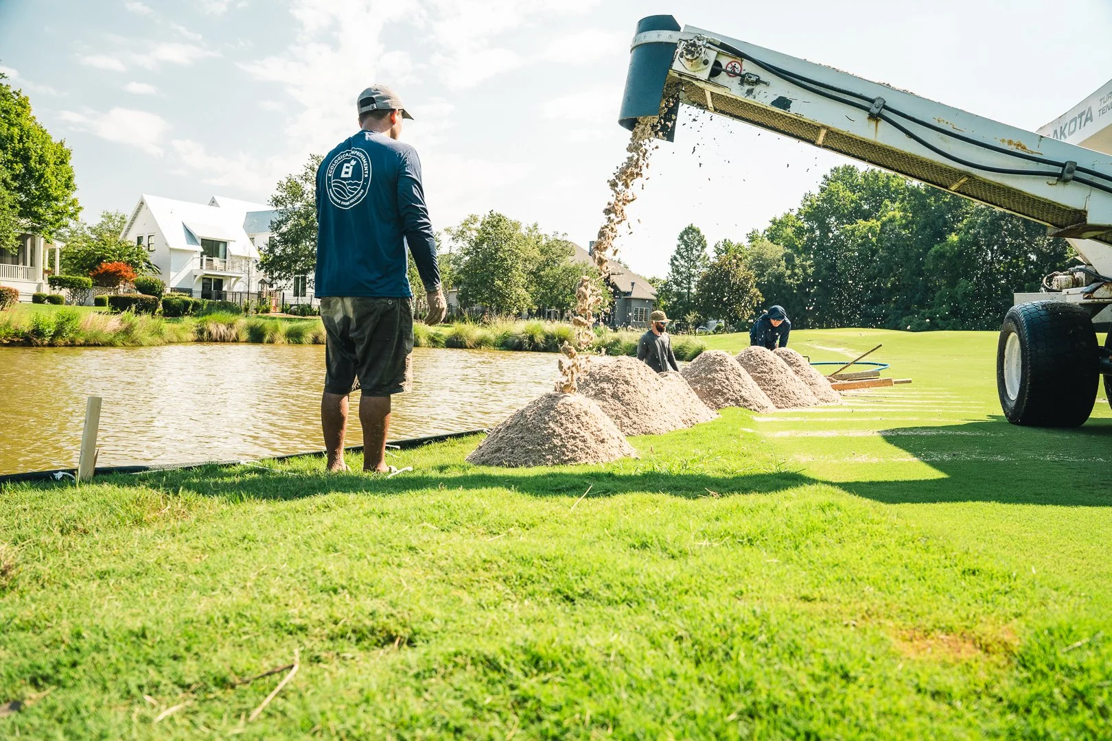 Workers using a sand and water mixture to clean or build a sandbag dam along a riverbank, with a large machine pouring the mixture over the sand.