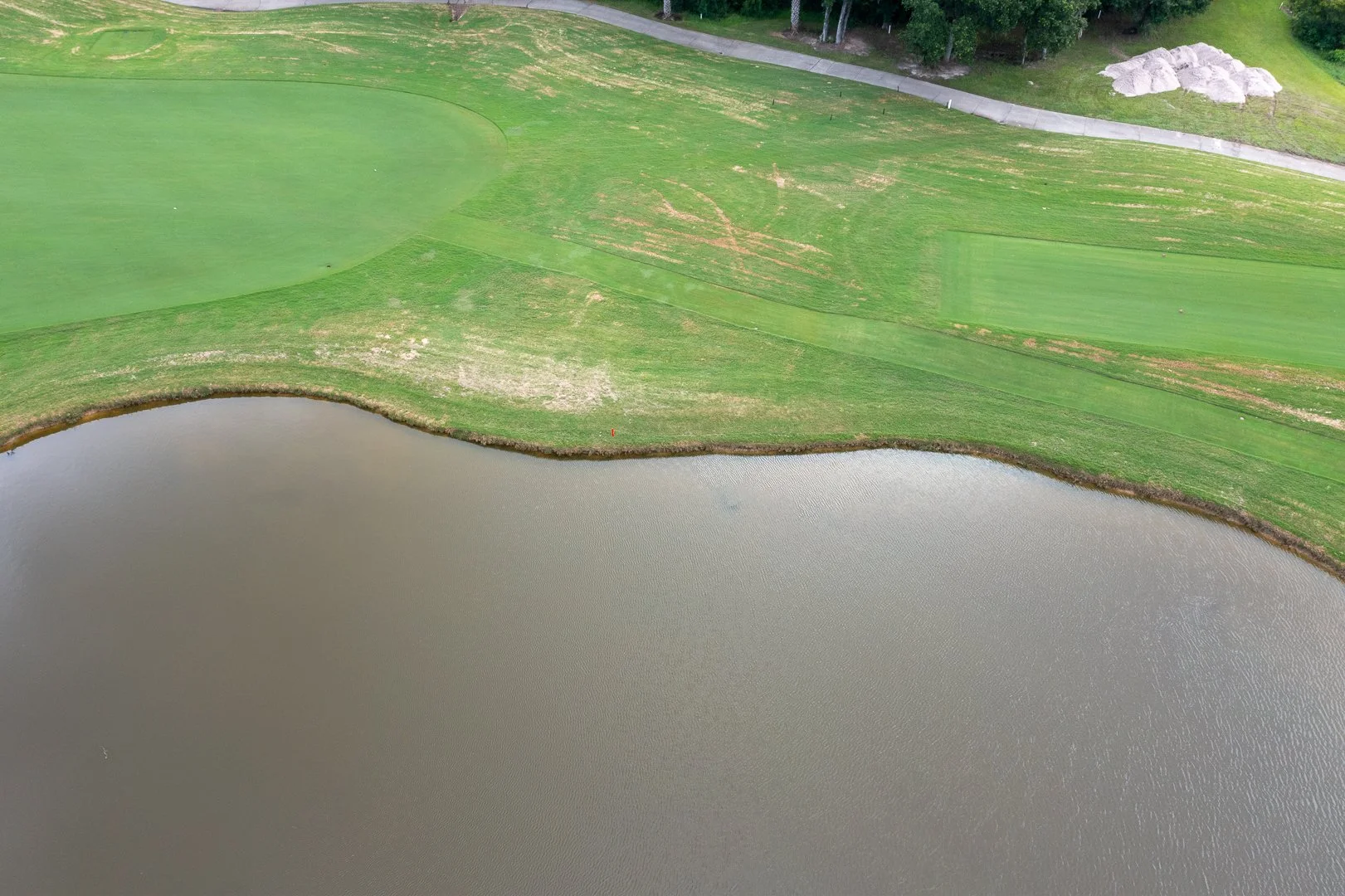 Aerial view of a golf course with green grass, a water hazard, a paved cart path, and some trees.