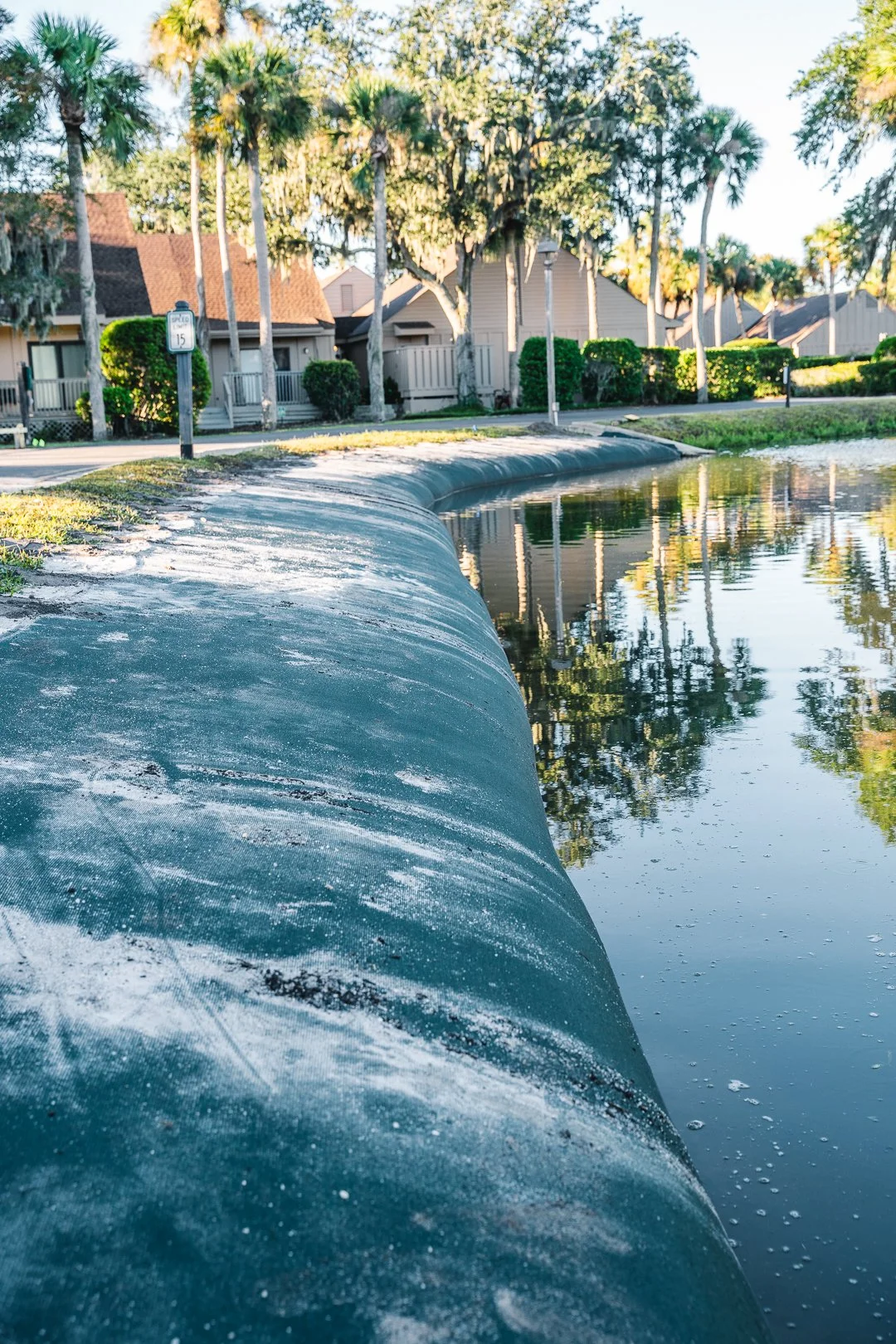 A canal lined with residential houses and palm trees, with a sidewalk on the side nearest the camera, covered in chalk or salt. The canal water is calm and reflects the trees and sky.