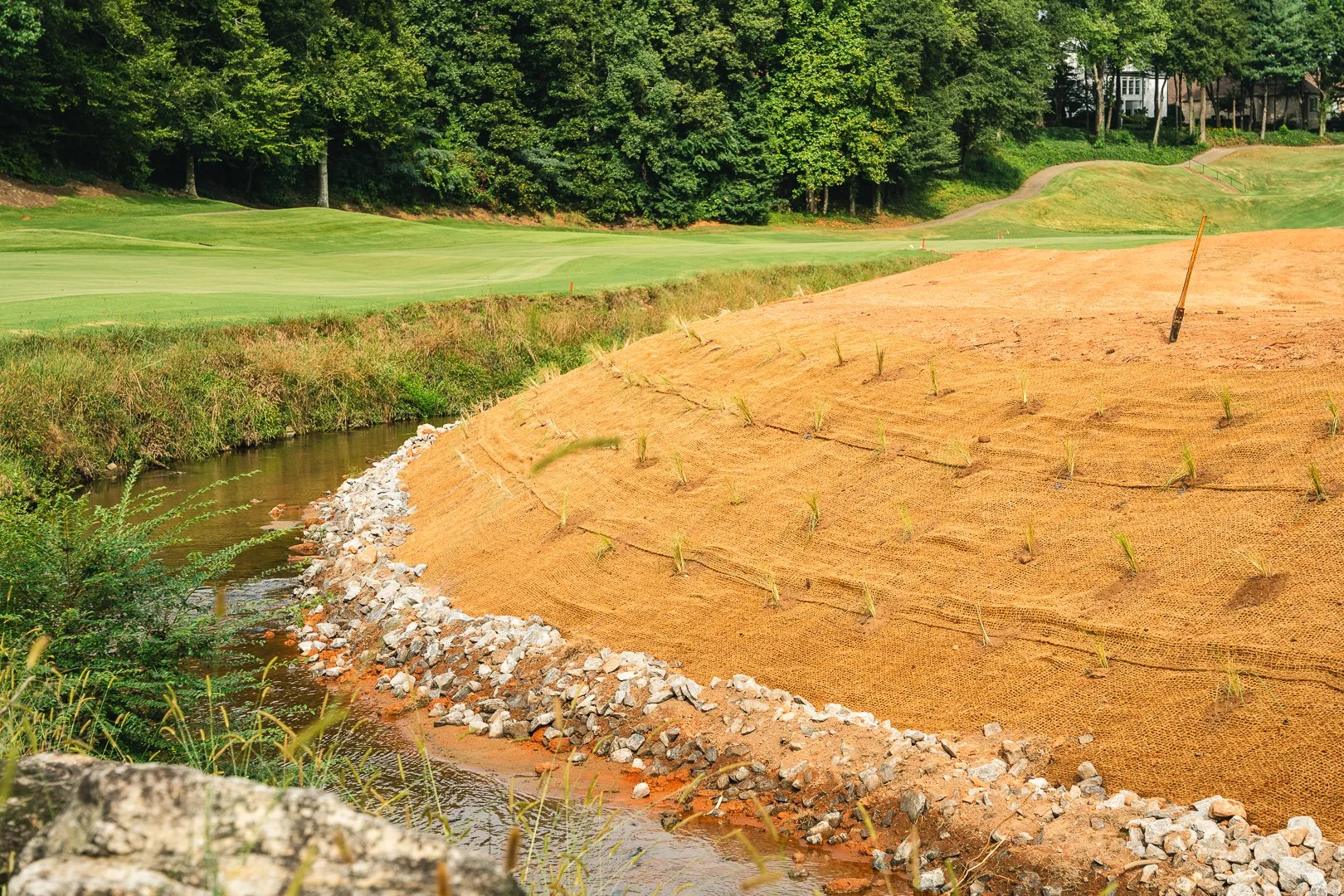 A golf course under construction with a newly seeded slope, a small stream, and green fairways in the background, surrounded by trees.