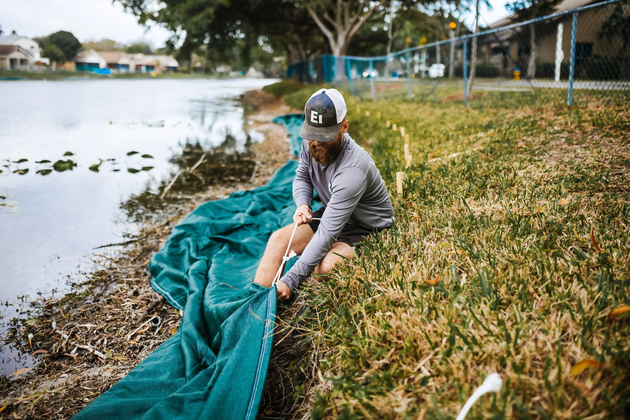 A man with a beard, wearing a gray jacket, black shorts, and a gray and black cap, is sitting on the grassy bank of a lake, securing a green fishing net along the shoreline. There are trees, houses, and a blue chain-link fence in the background.