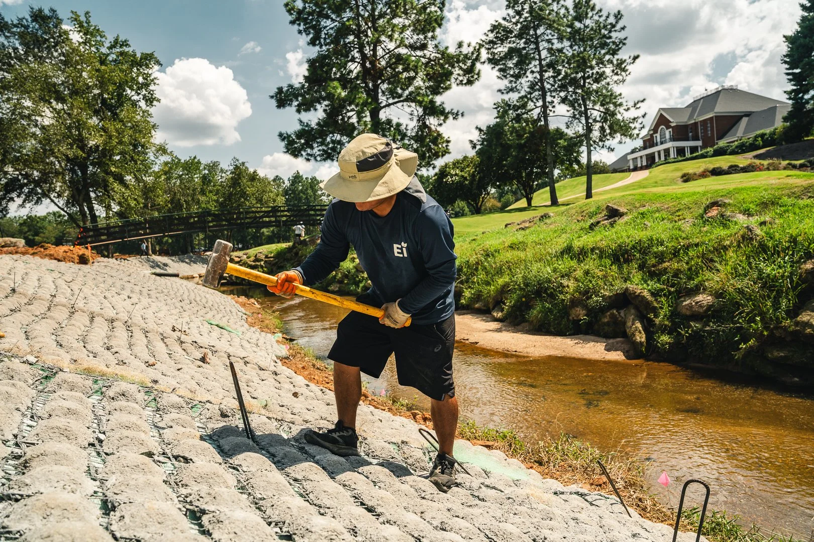 A construction worker paving a pathway with bricks near a small stream in a park-like area.