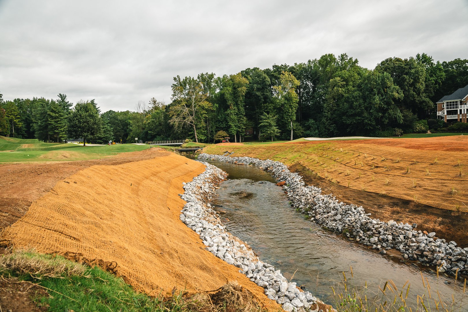 A small stream flowing through a landscaped area with some construction materials and orange netting on either side, in a suburban neighborhood with trees and houses in the background under an overcast sky.