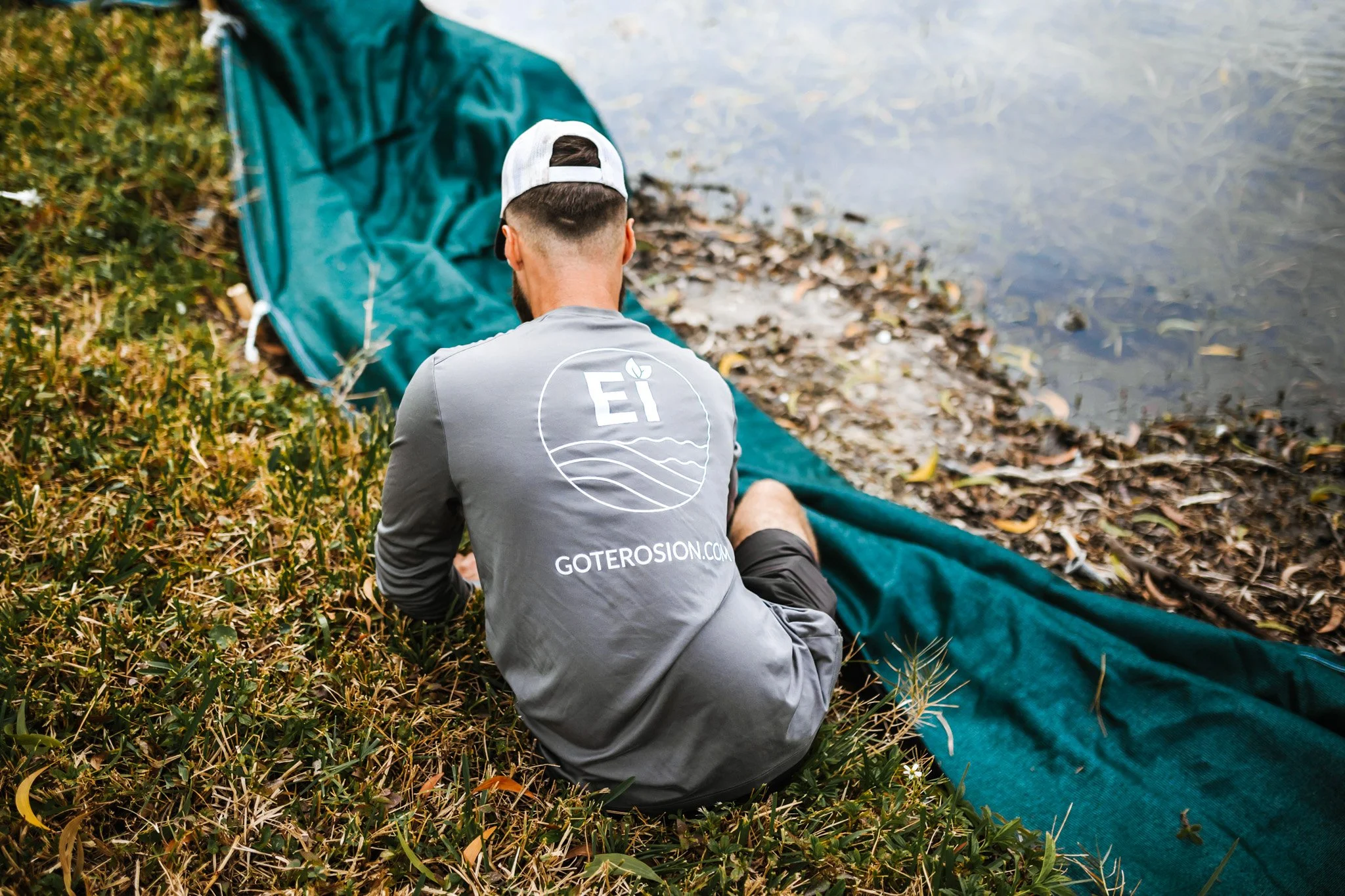 A man sitting on a grassy bank by a lake, facing away, wearing a gray shirt with 'GOTORosion.com' and a logo on the back, a white cap, and shorts, with a blue fishing net or barrier set up by the water.