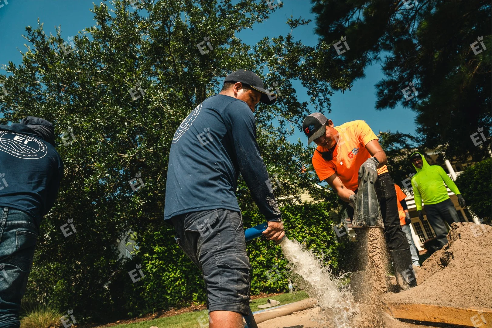 Group of people working on a construction site, with two men handling a water hose and pipe, surrounded by trees and a building in the background.