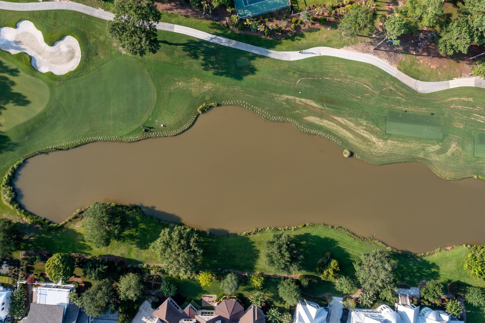 Aerial view of a park with a pond at the center, surrounded by grass, trees, walking paths, and residential houses.