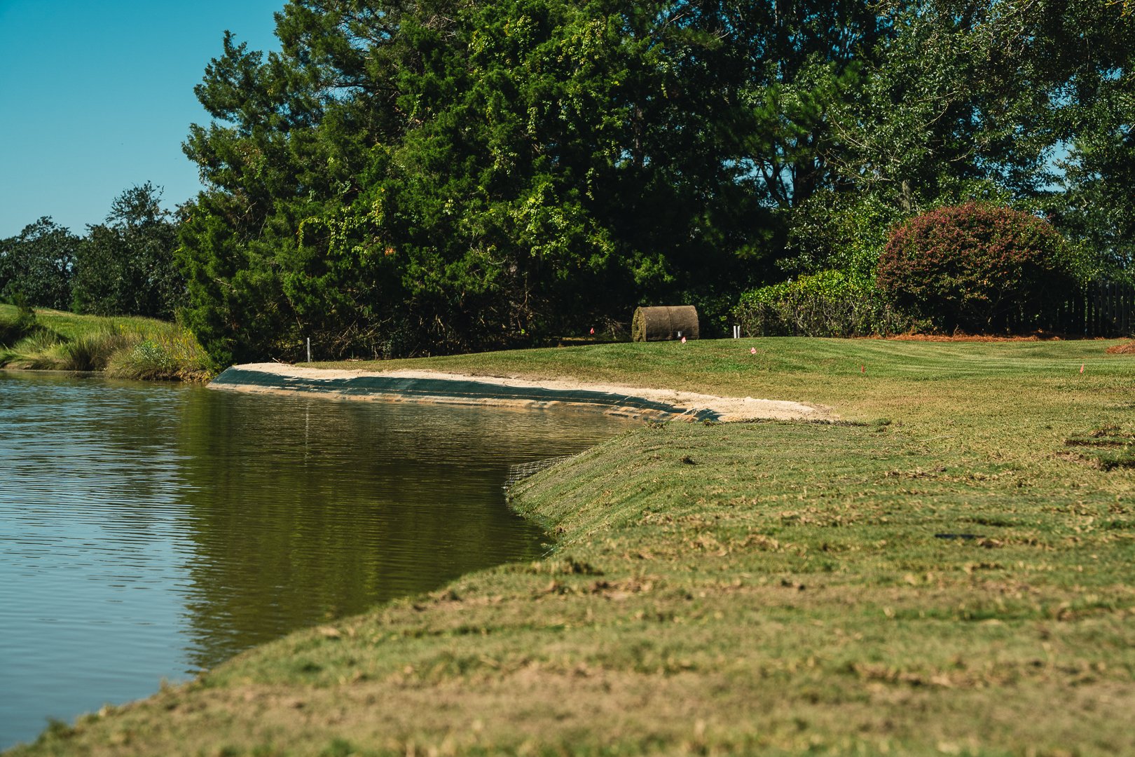 A lakeshore with a strip of grass and sand, trees in the background, and calm water reflecting the sky.