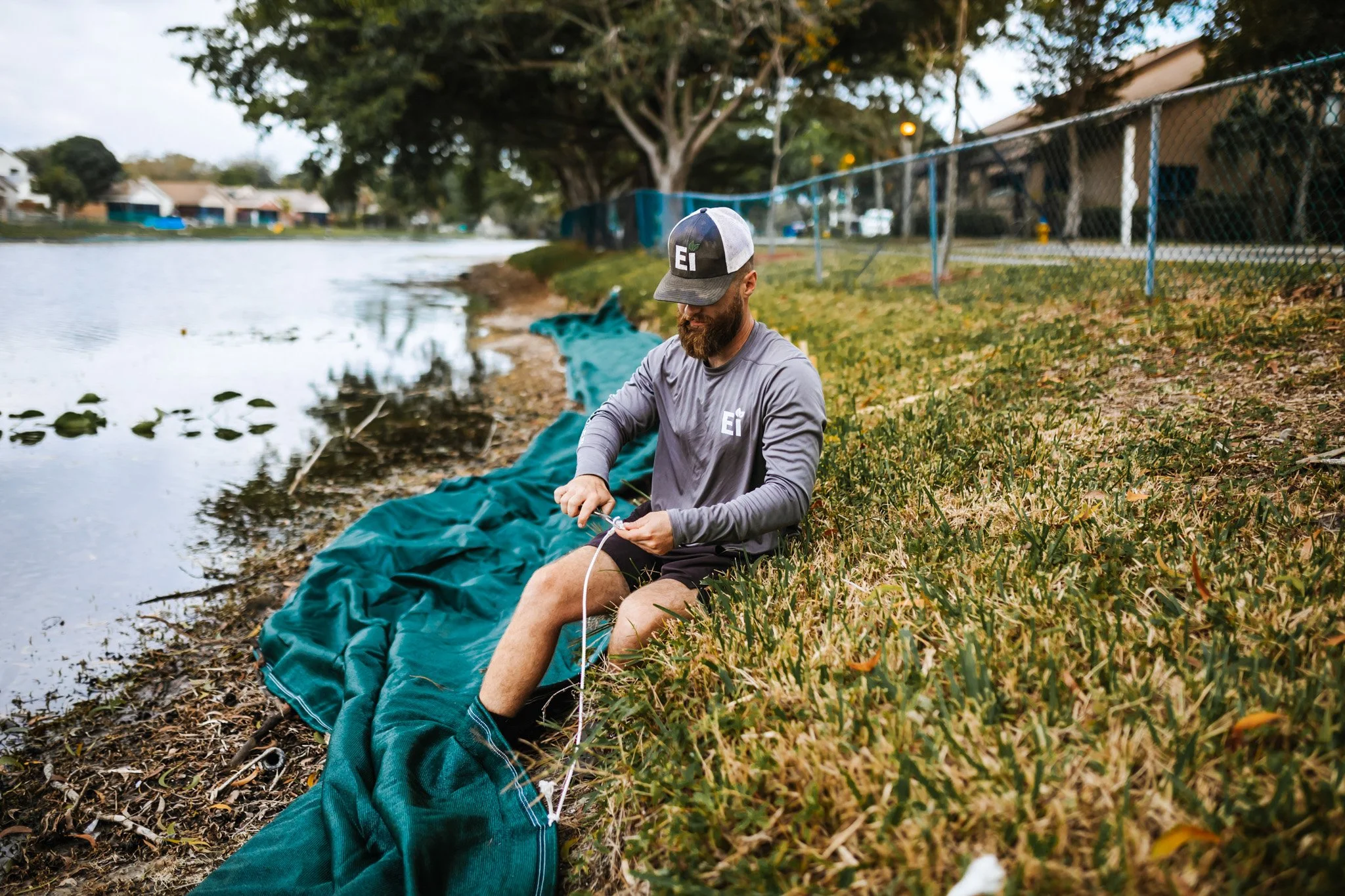 Man sitting by a lake, preparing fishing gear, wearing a gray long sleeve shirt, black shorts, a cap, and socks, with a green fishing net on the ground near him.