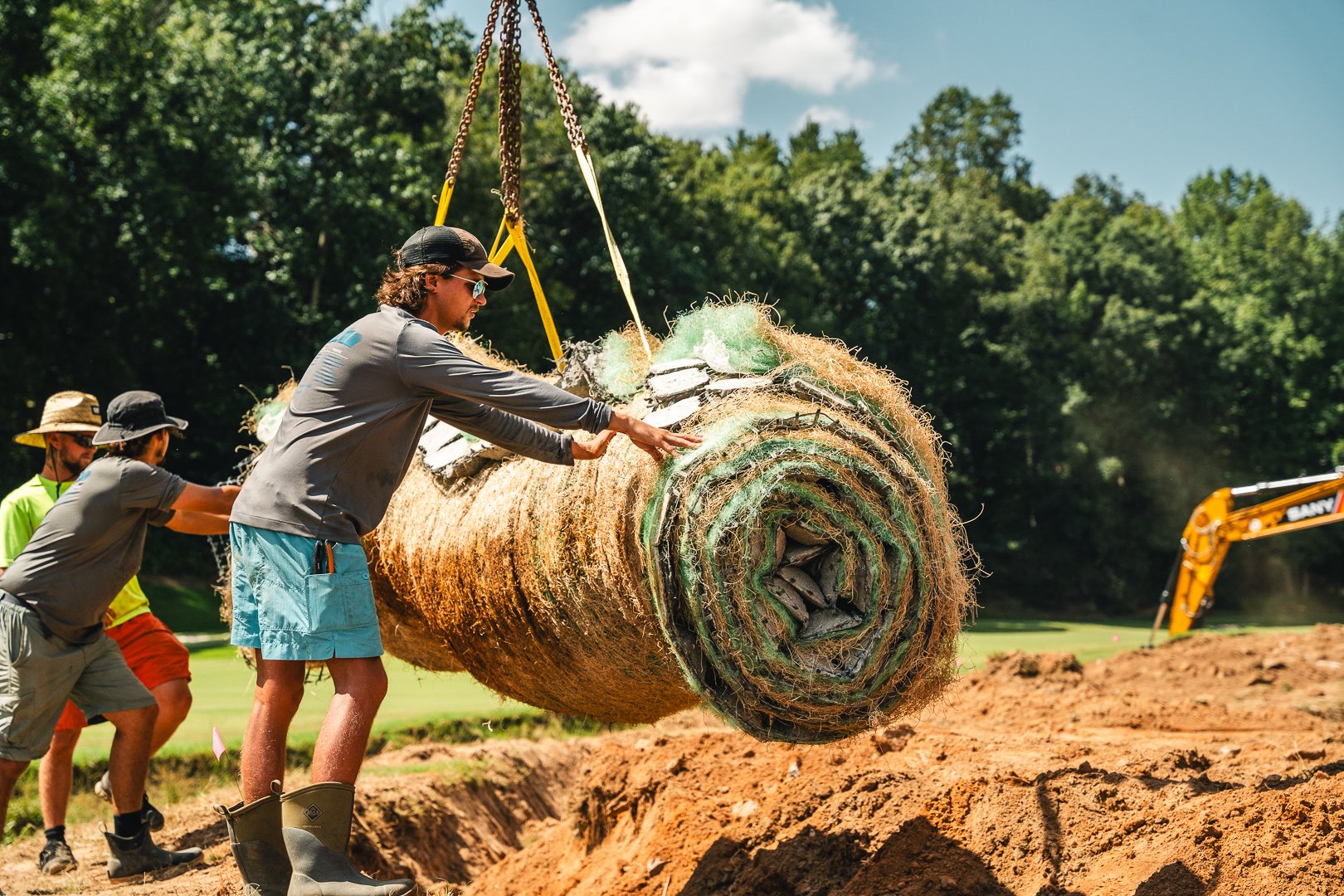 People working together to lift and position a large rolled-up turf roll on a construction site with a wooded background.