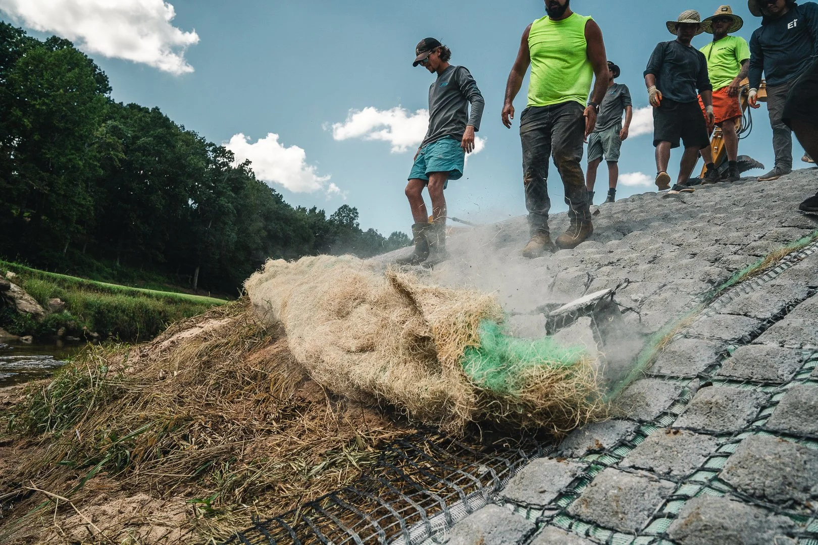 Group of people working on erosion control on a riverbank with gabion baskets, some wearing hats and boots, under a partly cloudy sky.