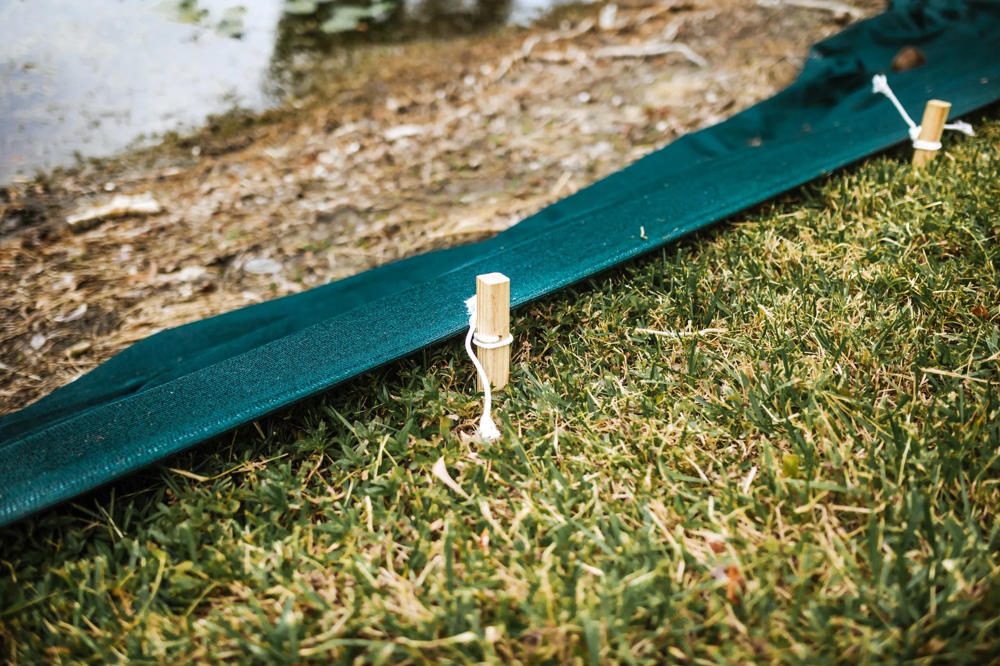 A close-up of a green fabric border held in place by wooden stakes with white ties in a grassy area near a body of water.