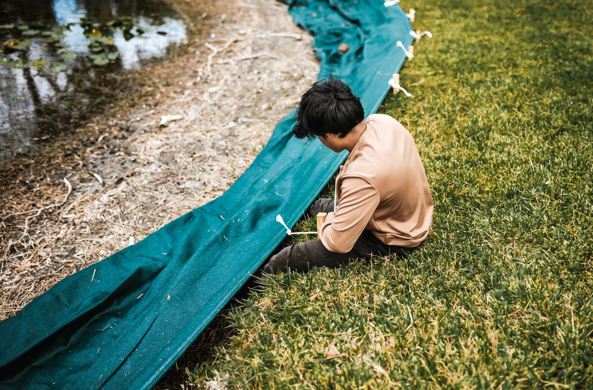 Person sitting on the grass near a pond, working on setting up a green water barrier or liner along the edge of the pond, surrounded by greenery.