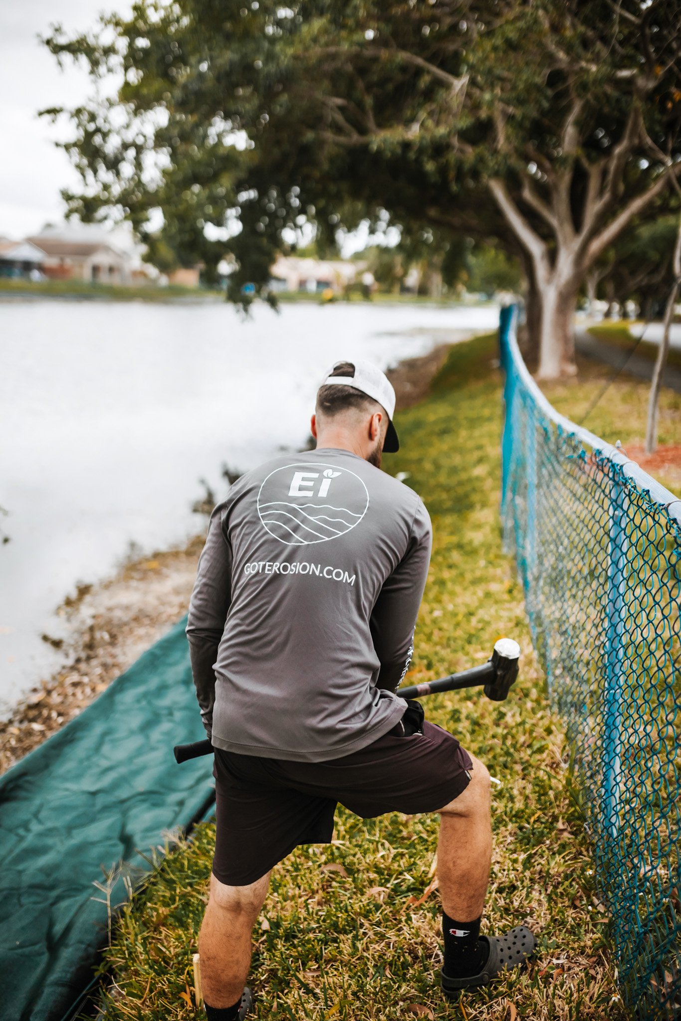 A man wearing a gray long-sleeve shirt, black shorts, black Crocs, and a white cap is using a hammer to plant something in the ground next to a blue safety fence beside a body of water with trees and houses in the background.
