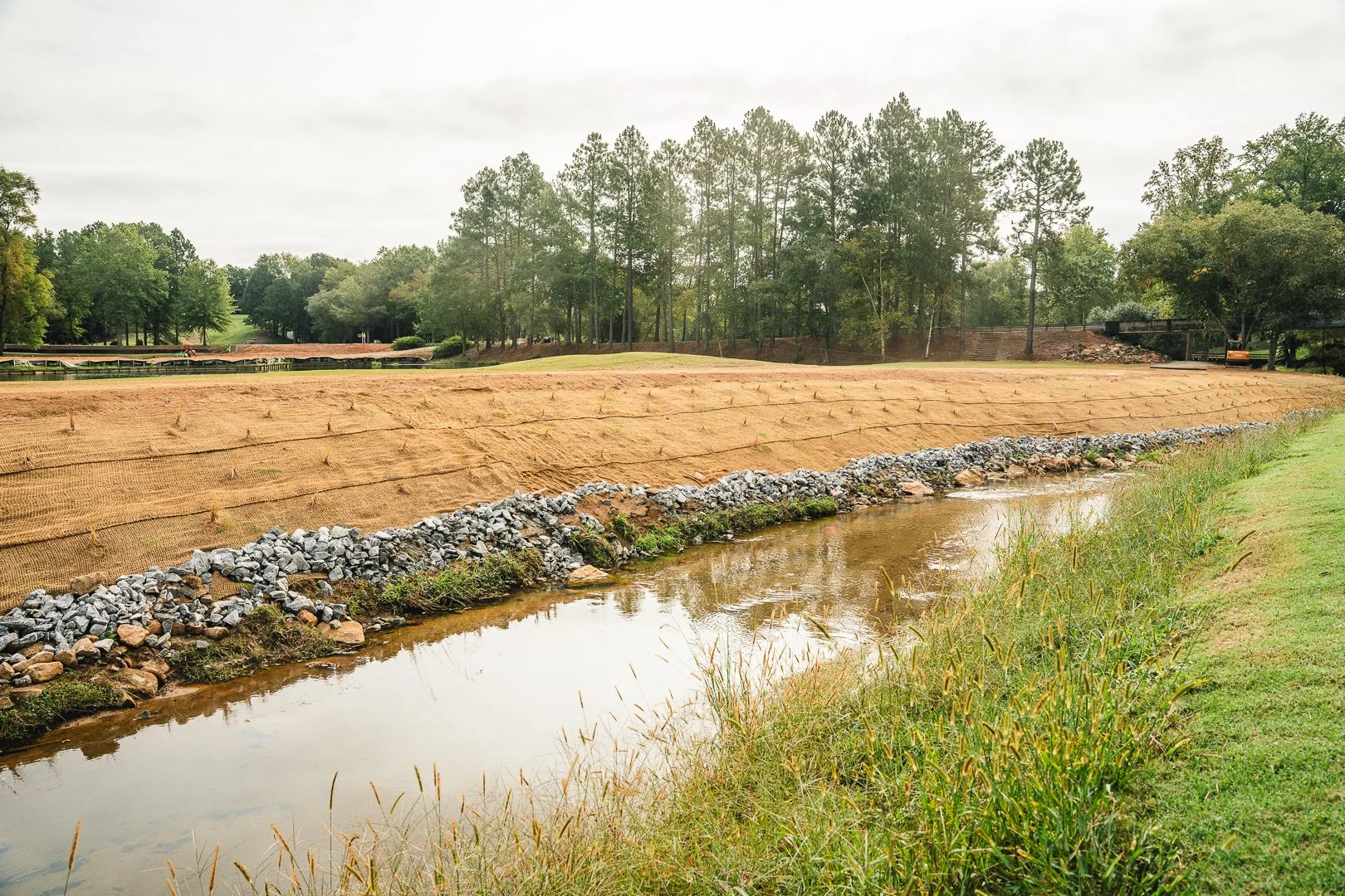 Construction site with a dirt area covered by protective fabric, a small stream with a rock border, and surrounded by trees under an overcast sky.