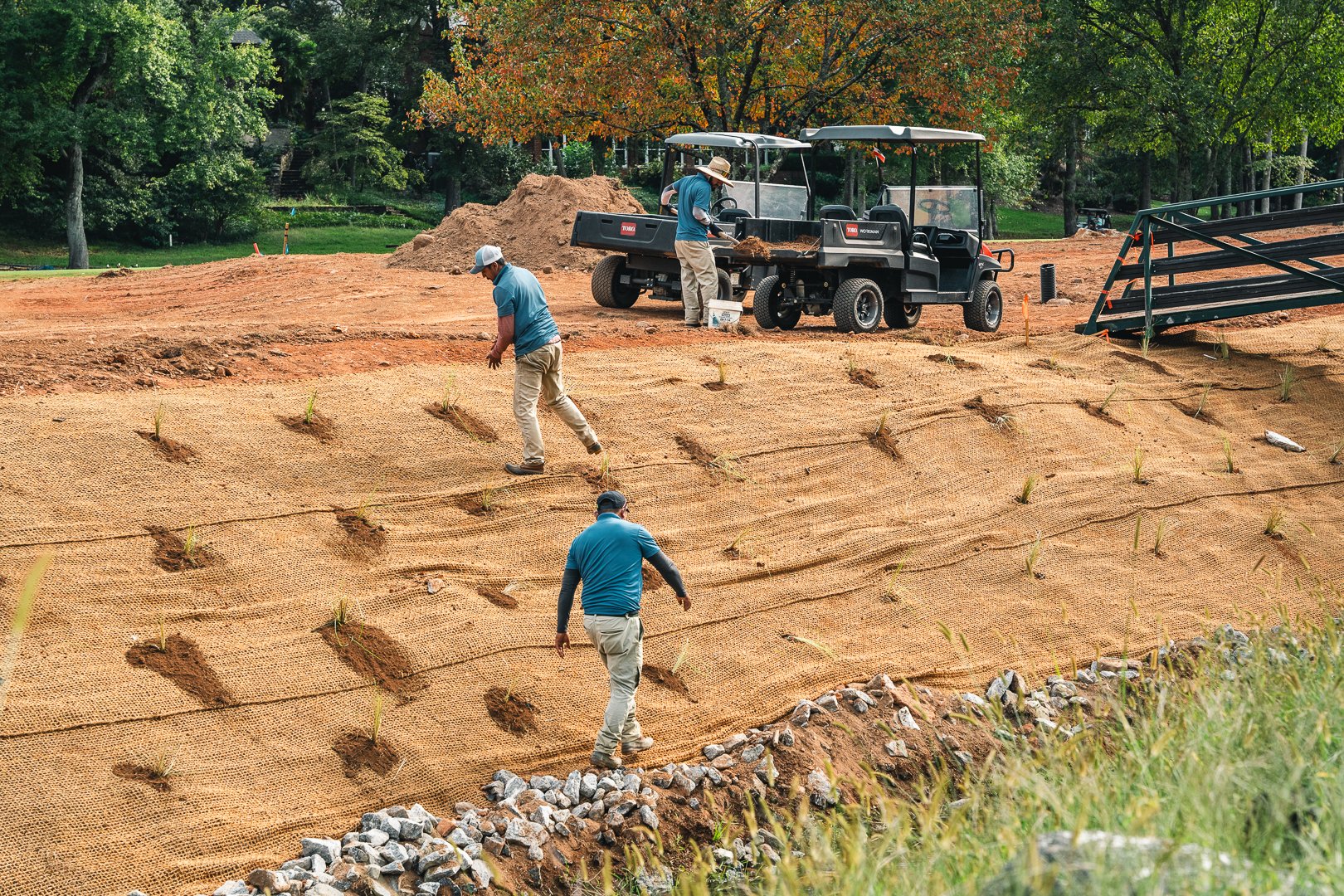 Three workers install erosion control fabric on a sloped landscape with small plants. A pickup truck and tools are nearby, surrounded by trees and natural scenery.