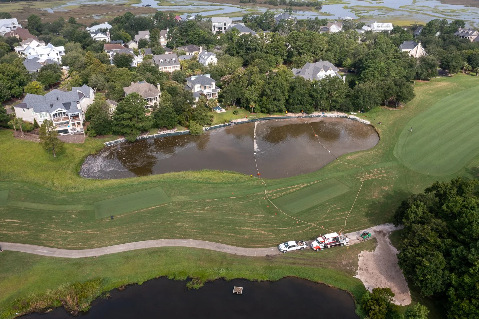 Aerial view of a residential neighborhood near a golf course with a water hazard. A water treatment or drainage process is being conducted in the pond, with equipment and vehicles parked nearby. Trees, houses, a walking path, and a golf green are visible.