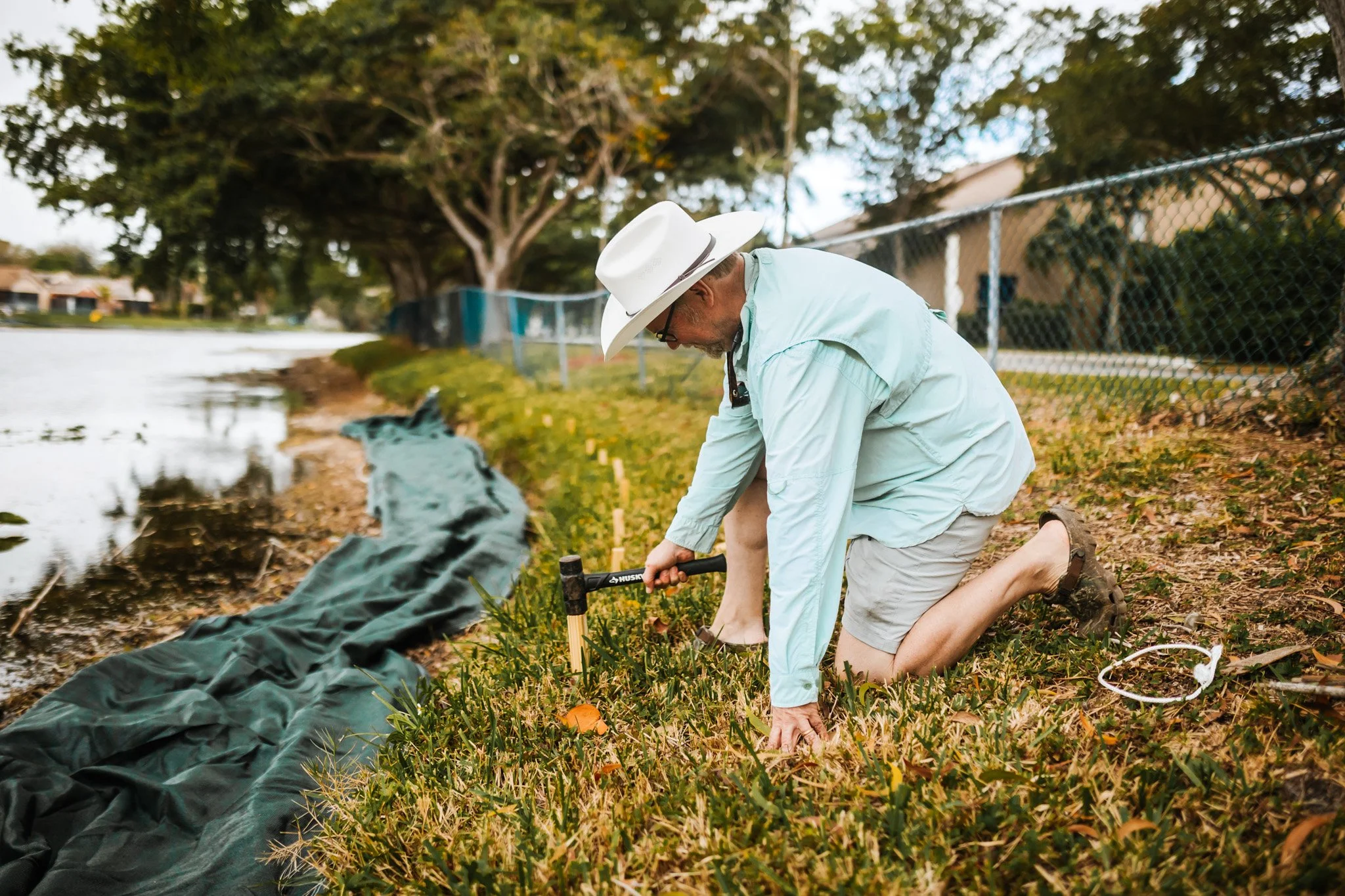 A man wearing a wide-brimmed white hat and beige shorts is kneeling on the grass near a body of water, using a gardening tool to plant or tend to plants along the shoreline, with a black tarp partially laid out behind him.