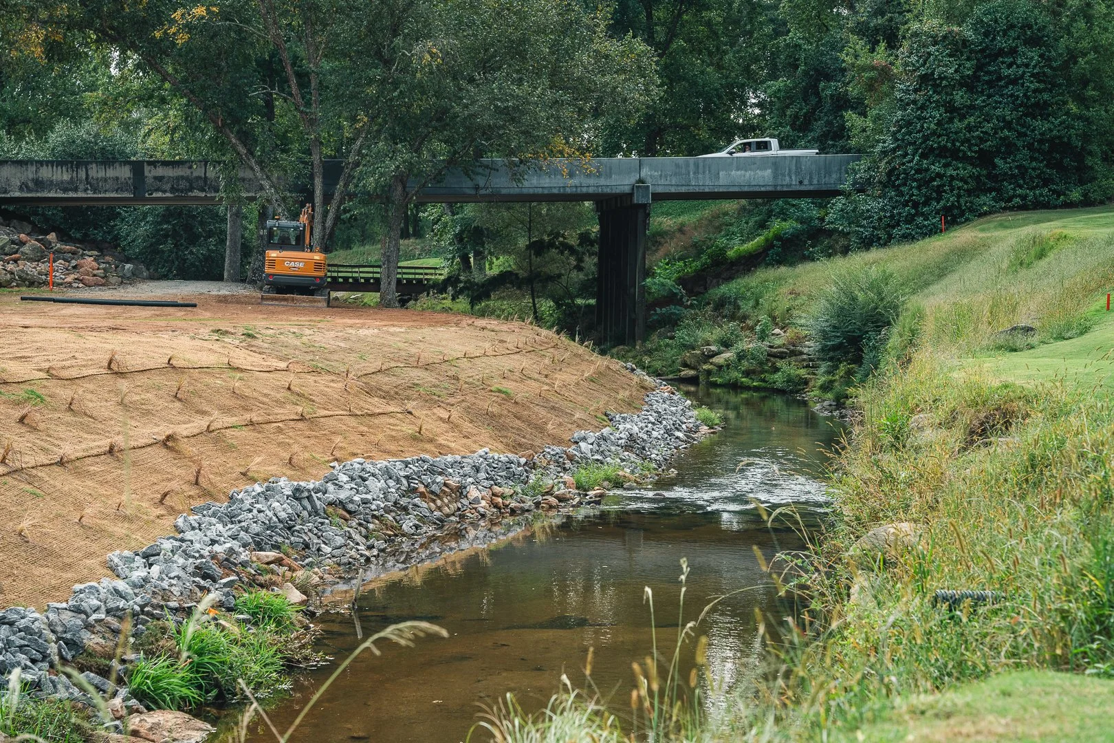 Construction site near a creek with a bridge and trees, featuring a small excavator and a car on the bridge.