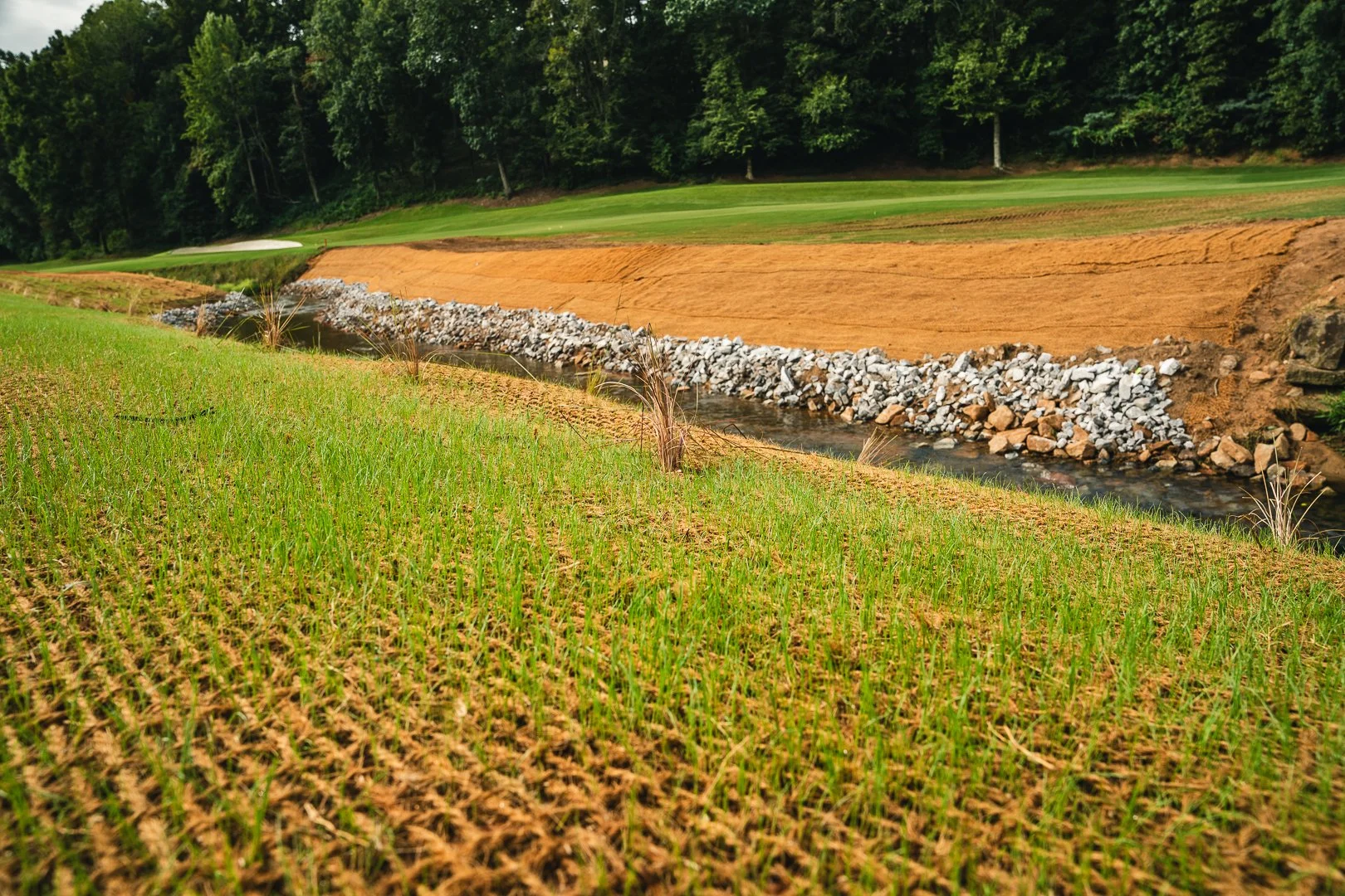 A golf course with a small stream running through it, surrounded by grassy areas and an orange dirt slope, with trees in the background.